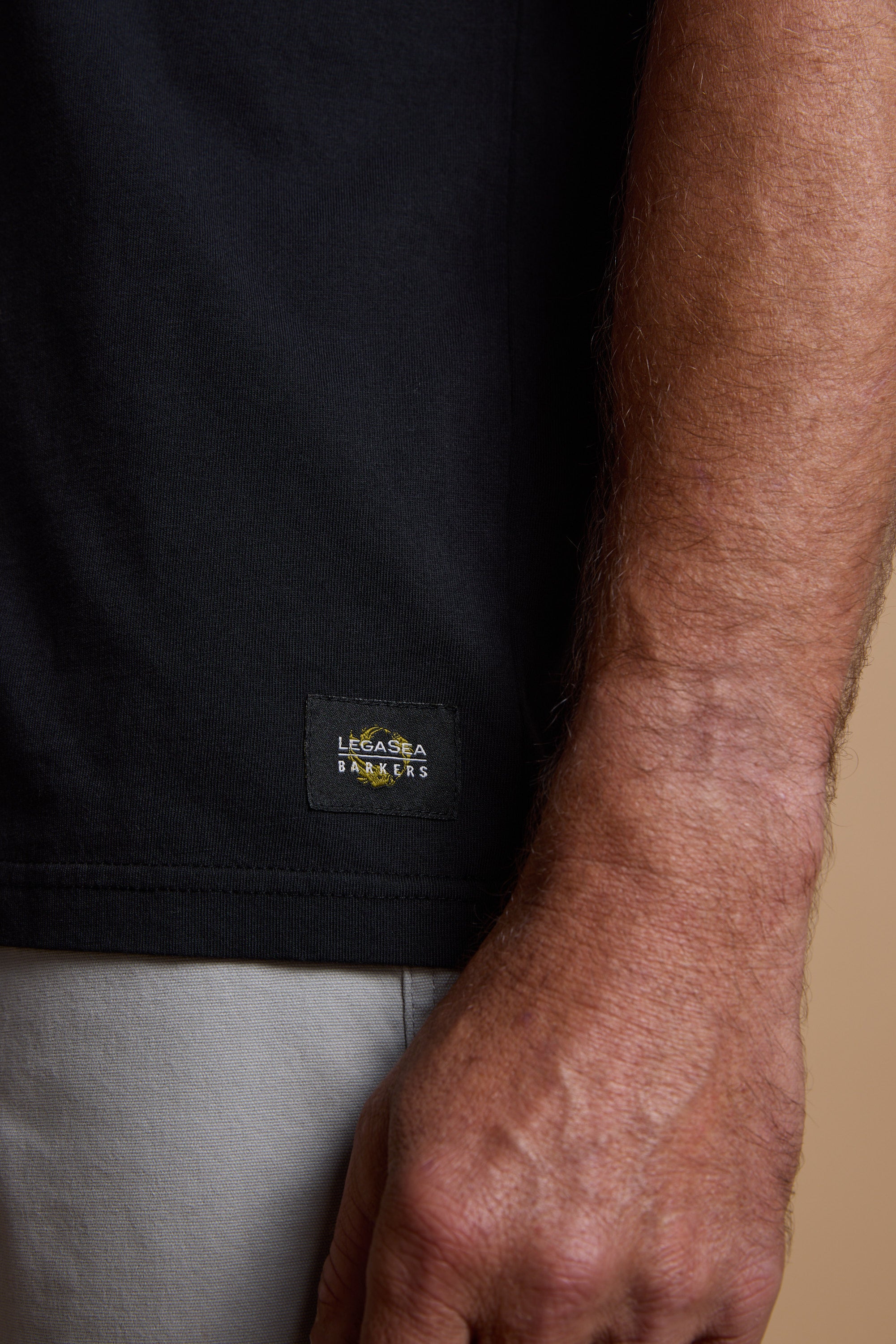 A close-up of a person's right arm and hand in light-colored pants features the Legasea Tail Print Tee by Legasea, a black organic cotton shirt with a small branded label near the hem.