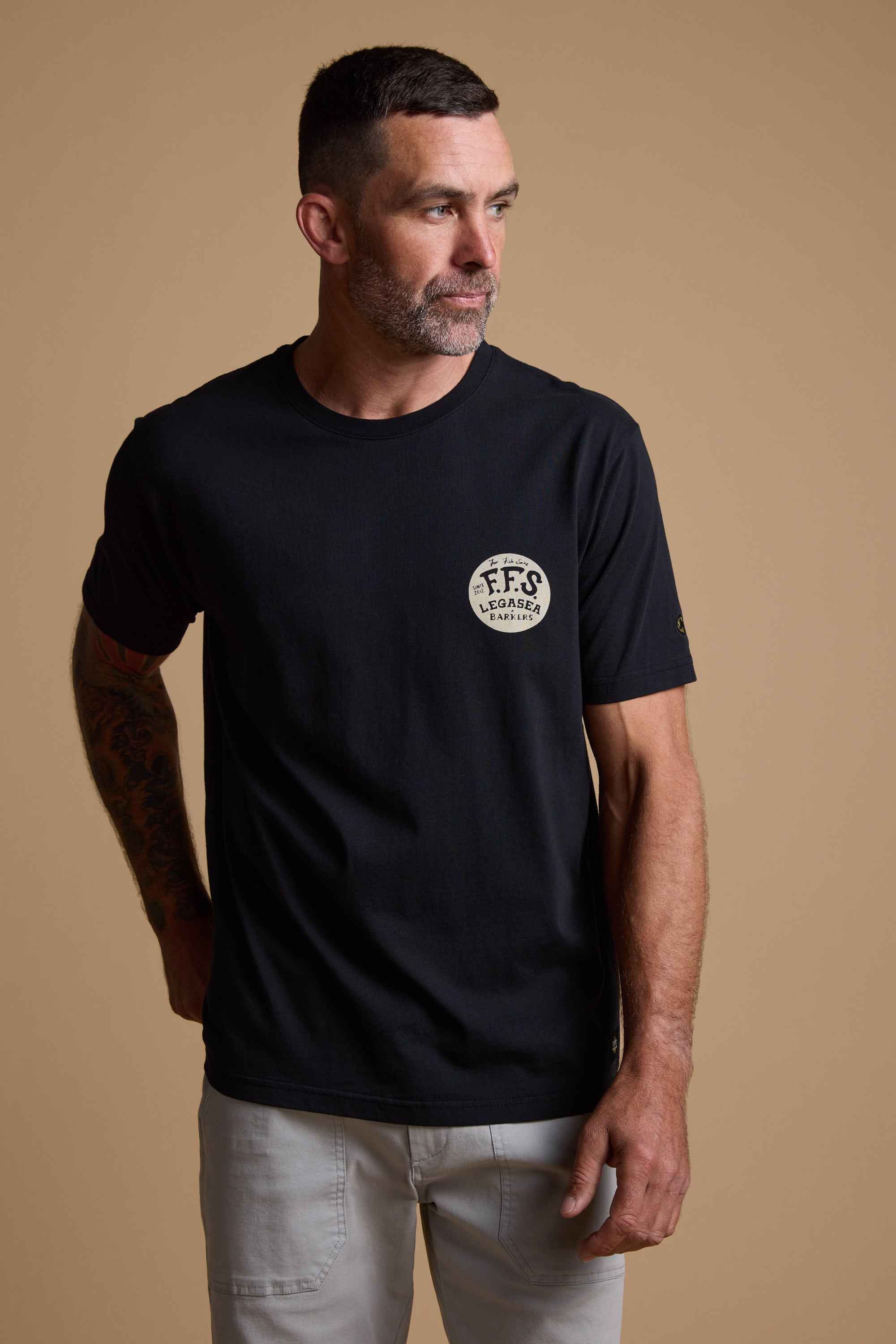 A man with short dark hair and a beard wears the Legasea Tail Print Tee—a black organic cotton T-shirt by Legasea with a small circular logo on the chest—and light-colored pants, standing against a plain beige background.