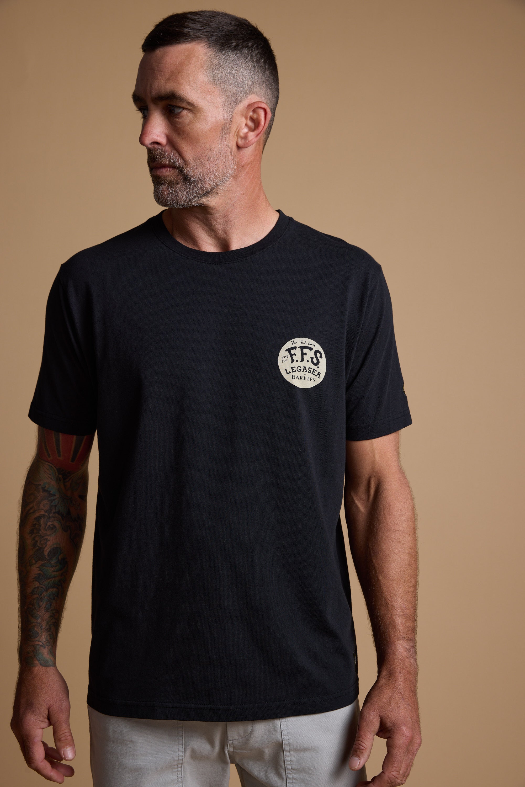 A man with short dark hair and a beard, wearing a black Legasea Tail Print Tee featuring a round “FFS Legends” logo on the chest, stands against a beige background, looking left and showing a tattoo on his right arm. Brand: Legasea.
