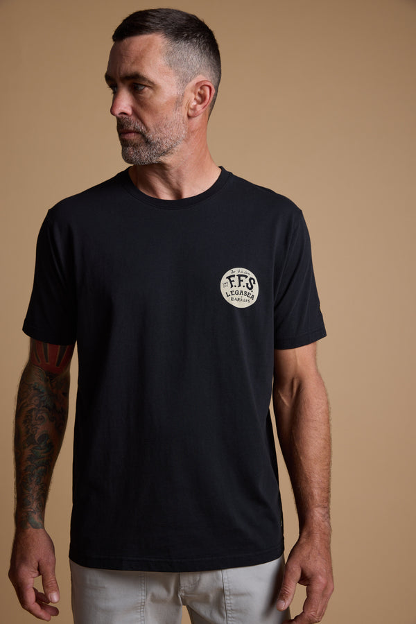 A man with short dark hair and a beard, wearing a black Legasea Tail Print Tee featuring a round “FFS Legends” logo on the chest, stands against a beige background, looking left and showing a tattoo on his right arm. Brand: Legasea.