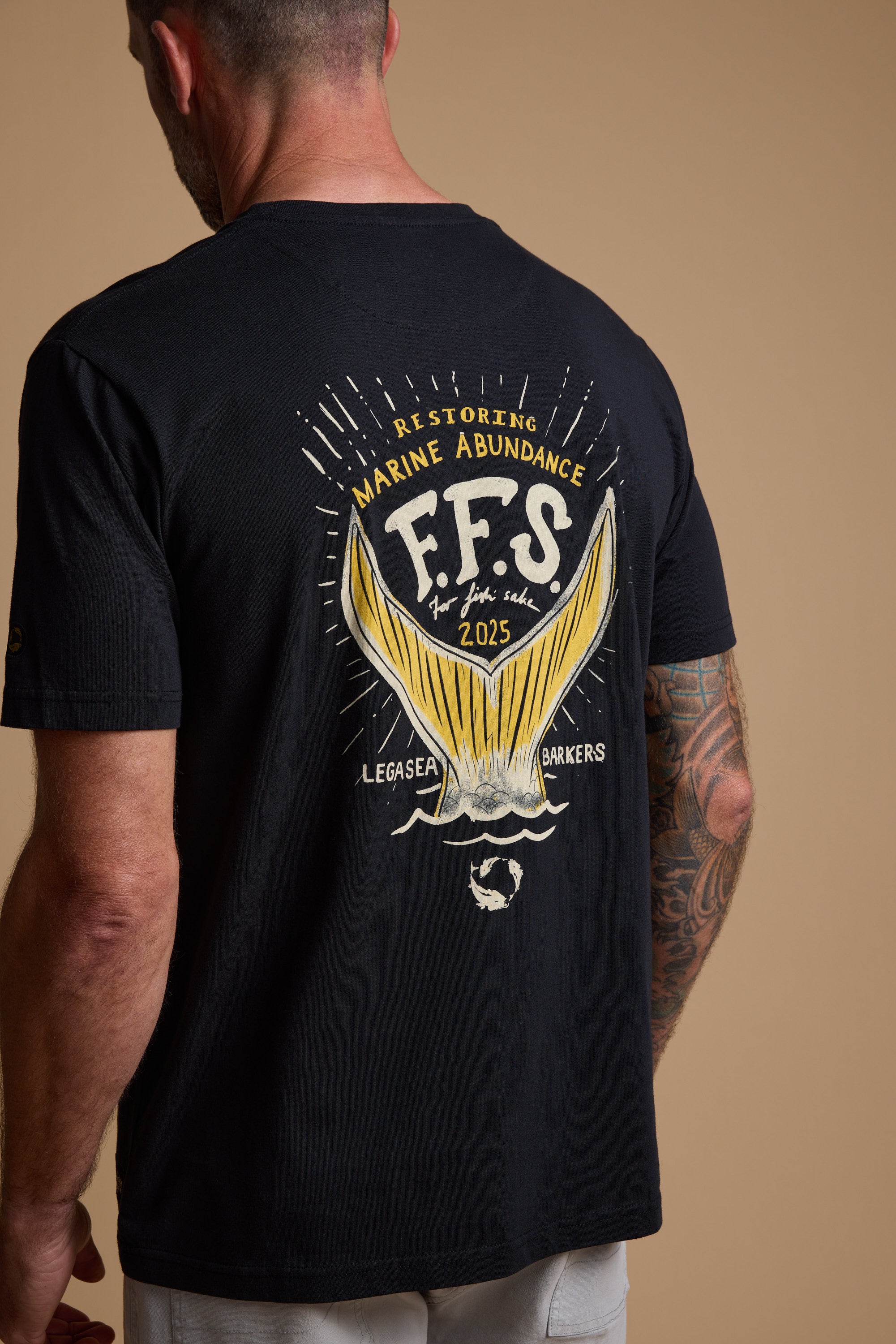 A man with a tattooed arm wears the organic cotton Legasea Tail Print Tee, featuring a yellow fish tail and "Restoring Marine Abundance, F.F.S. For fish sake, 2025, Legasea Barkers"—ideal for New Zealand coastline lovers.