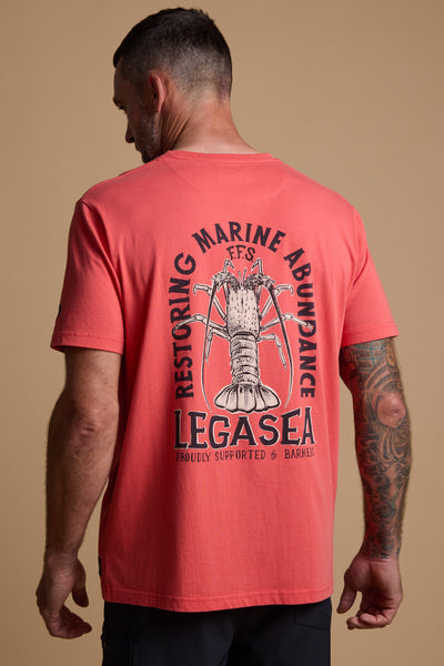 A man with short hair and a tattooed arm wears the Legasea Crayfish Print Tee in coral, featuring a bold crayfish graphic and celebrating New Zealand’s coastline, from the Legasea brand.