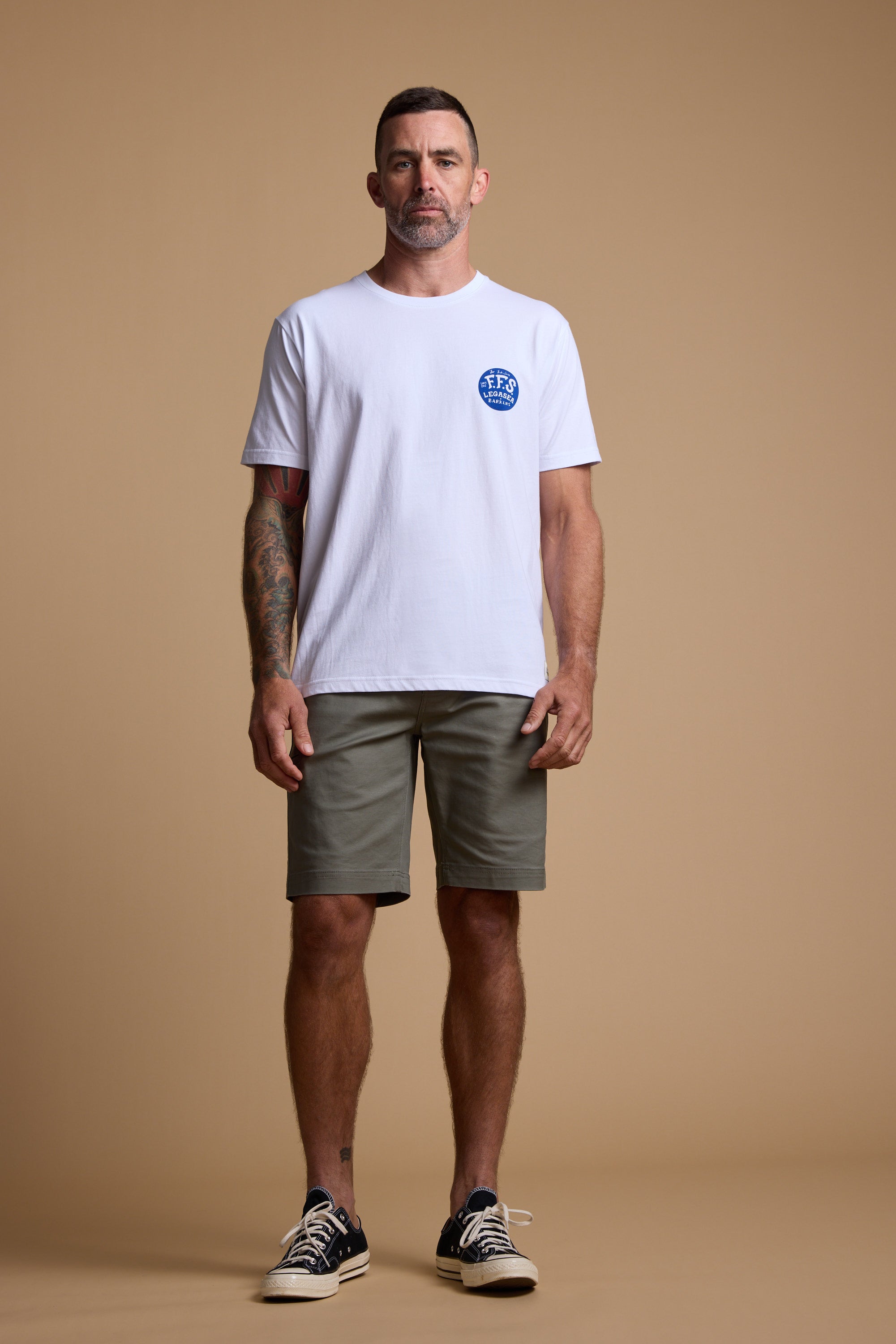 A man stands against a plain beige background, wearing the Legasea Marlin Print organic cotton tee with a blue circular logo on the left chest, olive green shorts, and black sneakers with white soles. One arm is tattooed as he looks ahead.