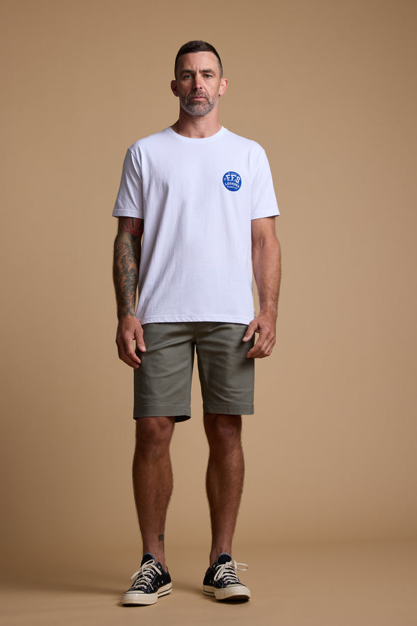 A man stands against a plain beige background, wearing the Legasea Marlin Print organic cotton tee with a blue circular logo on the left chest, olive green shorts, and black sneakers with white soles. One arm is tattooed as he looks ahead.