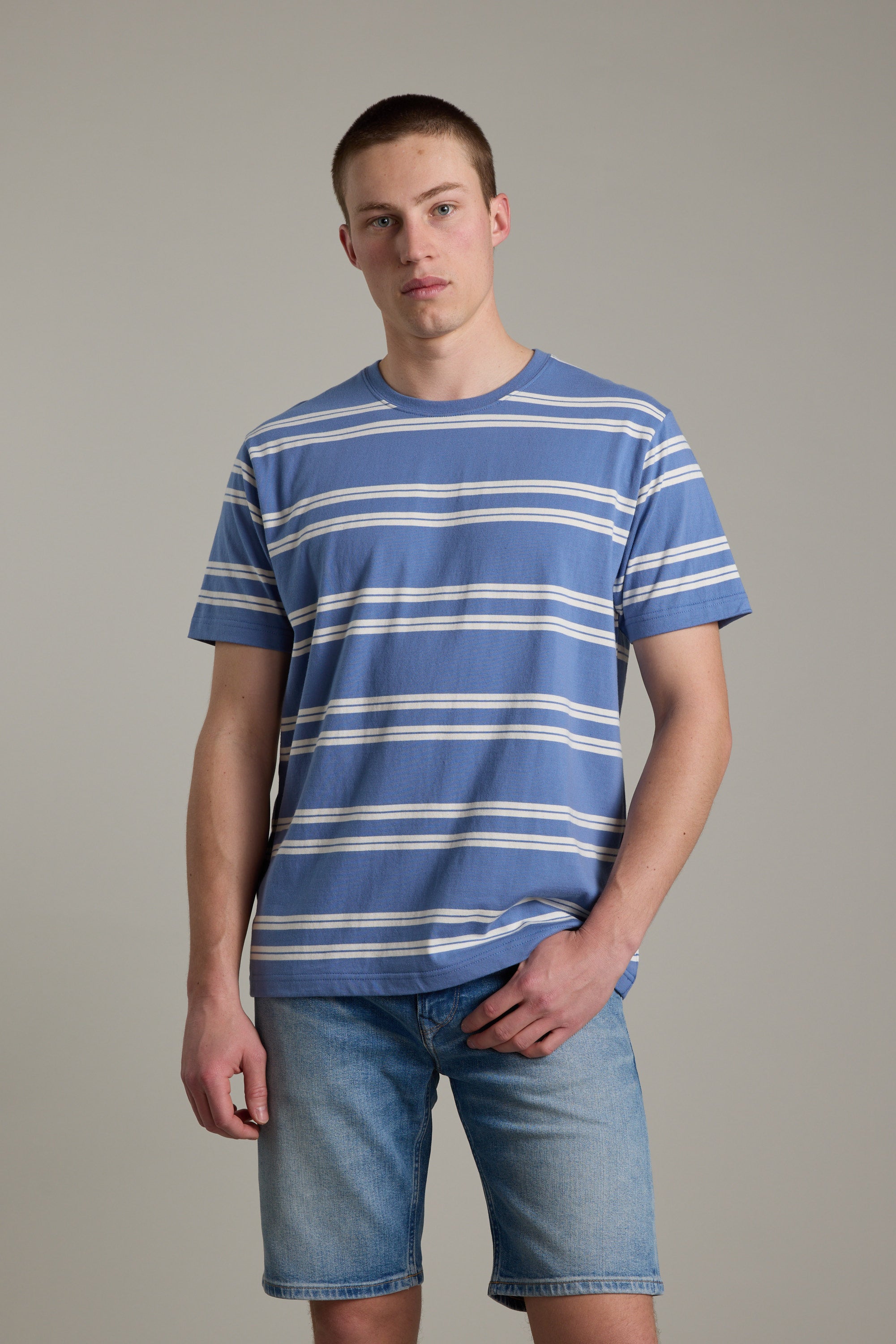 A young man with short hair and a neutral expression stands against a plain background, wearing the Barkers Northway Stripe Tee—an organic cotton blue-striped t-shirt—and light denim shorts, with one hand casually in his pocket.