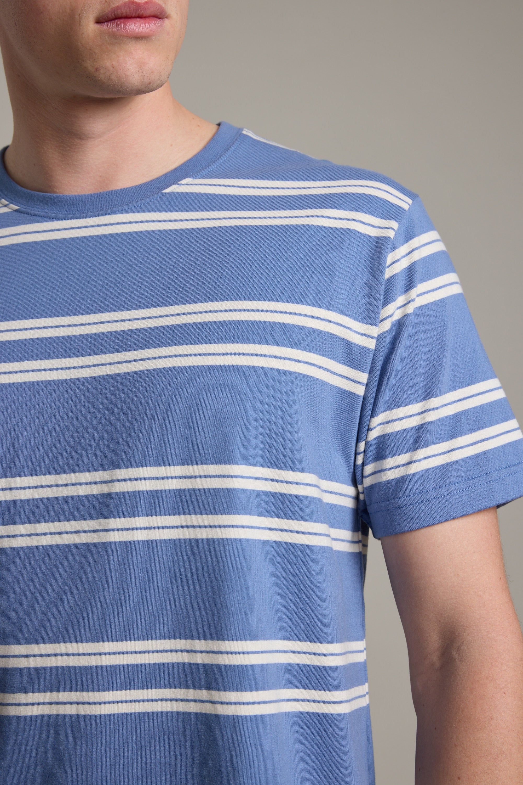 A person wears the Barkers Northway Stripe Tee, featuring blue and white horizontal stripes, shown from shoulders to above the elbow against a plain background with a partially visible face—a timeless summer essential.