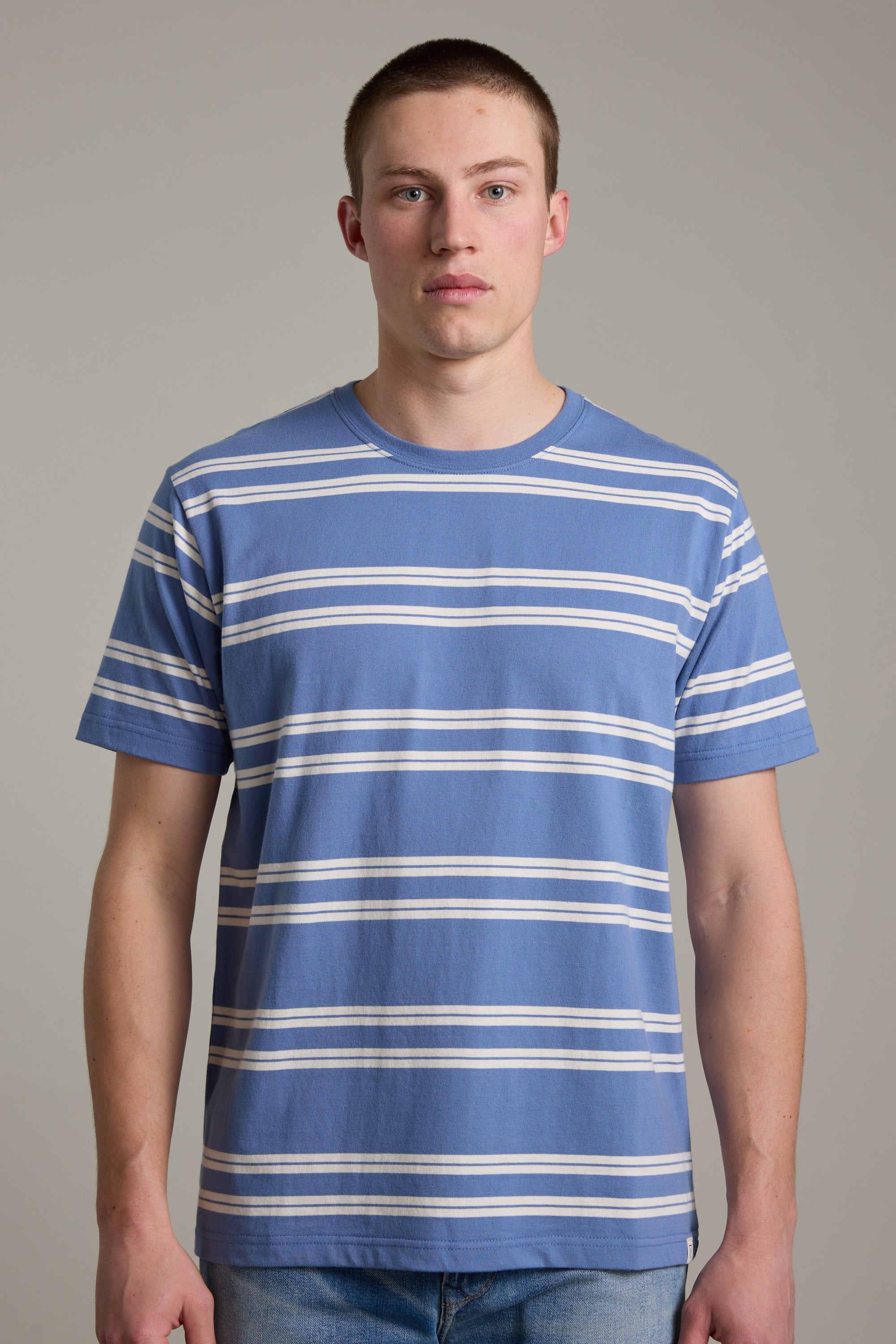 A young man with short hair wears the Barkers Northway Stripe Tee, featuring blue and white stripes, paired with light blue jeans against a plain light gray background—ideal for your summer wardrobe.