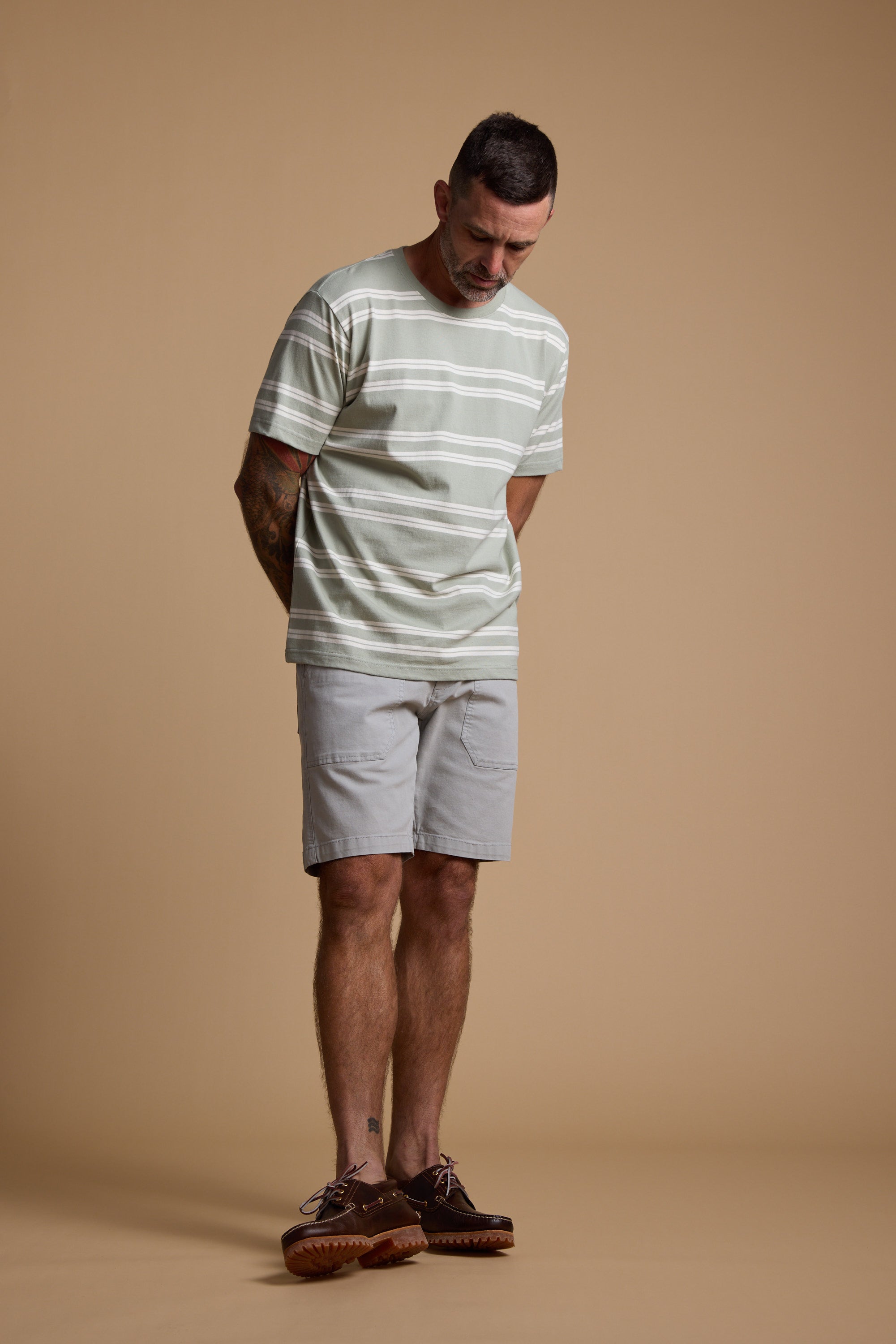 A man wears the Barkers Northway Stripe Tee in pale green and white, paired with light gray shorts and brown shoes, standing against a beige background—a relaxed choice for your summer wardrobe.