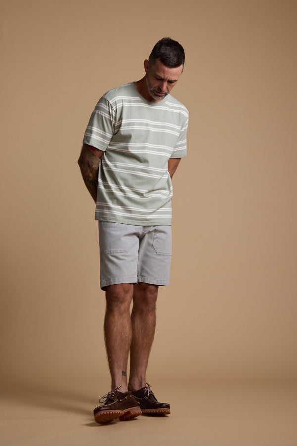 A man wears the Barkers Northway Stripe Tee in pale green and white, paired with light gray shorts and brown shoes, standing against a beige background—a relaxed choice for your summer wardrobe.