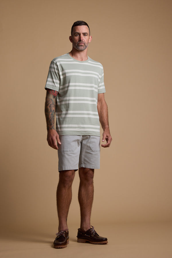 A man with short dark hair and a trimmed beard stands against a beige background, wearing Barkers' Northway Stripe Tee, light gray shorts, and brown shoes. One arm displays a colorful tattoo—his summer wardrobe essential.