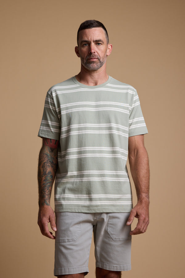 A man with short hair and a beard stands against a beige background, wearing the Barkers Northway Stripe Tee in light green and white stripes—an ideal summer choice. He has a tattoo on his left forearm and looks straight at the camera.
