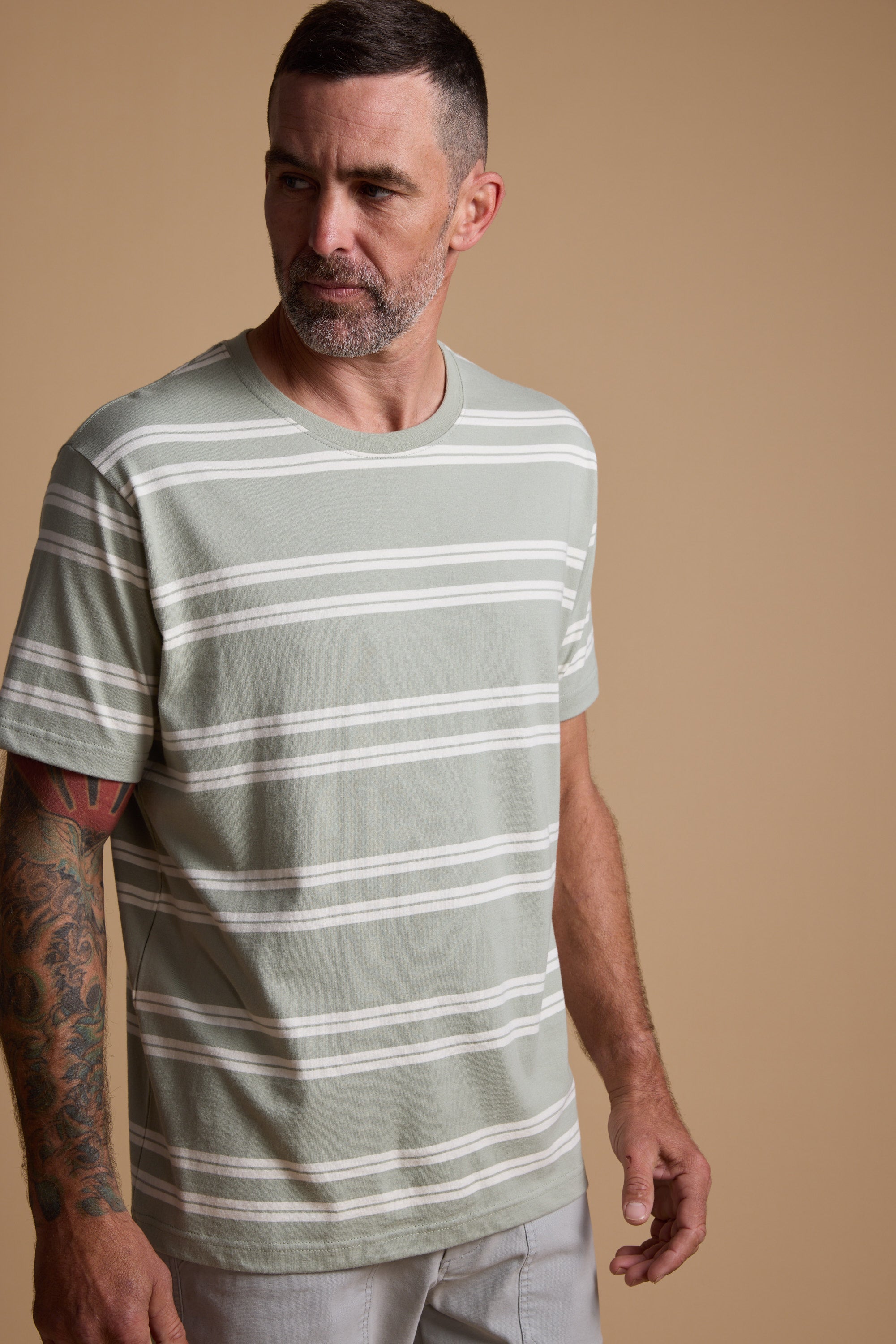 A man with short dark hair and a beard, wearing the Barkers Northway Stripe Tee made from organic cotton in light green and white, stands against a beige background. He has tattoos on his left arm—ideal for summer style.