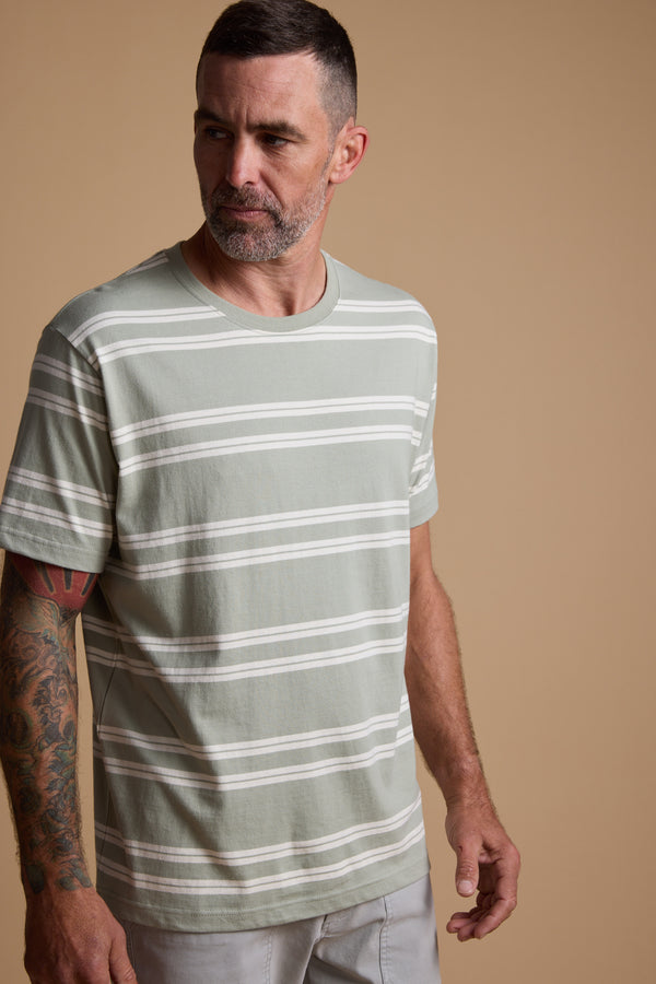 A man with short dark hair and a beard, wearing the Barkers Northway Stripe Tee made from organic cotton in light green and white, stands against a beige background. He has tattoos on his left arm—ideal for summer style.