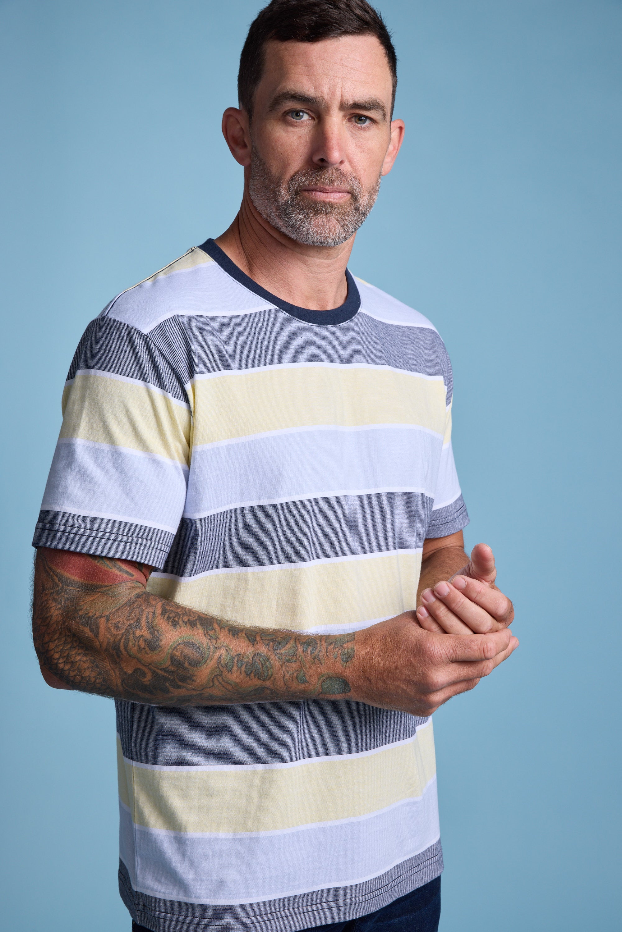 A man with short dark hair, a trimmed beard, and a tattooed left arm wears the Barkers Asbrooke Stripe Tee in gray, white, and pale yellow—its regular fit ideal for summer—standing against a plain blue background.