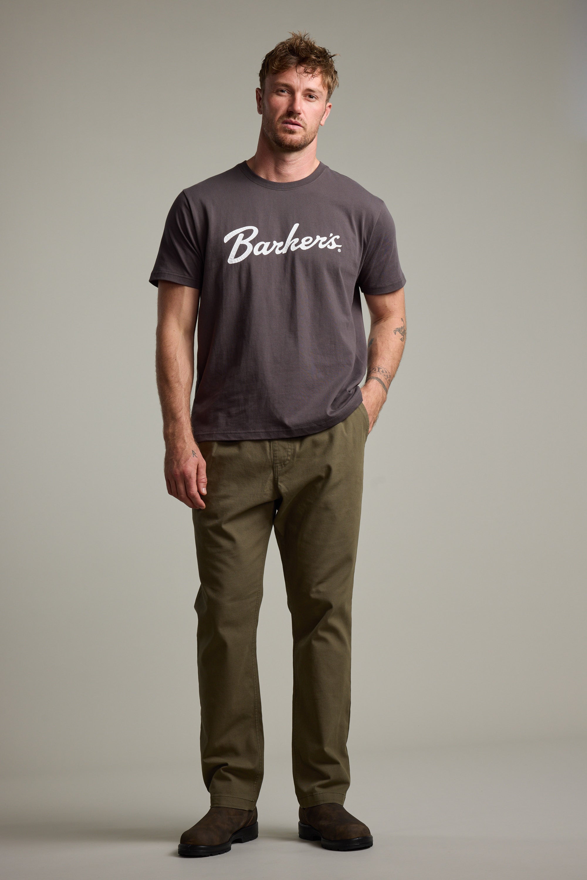A man with short light brown hair poses against a plain background, wearing a Barkers Finely Tuned Print Tee in grey, paired with olive green pants and brown suede shoes. He stands relaxed with one hand by his side.