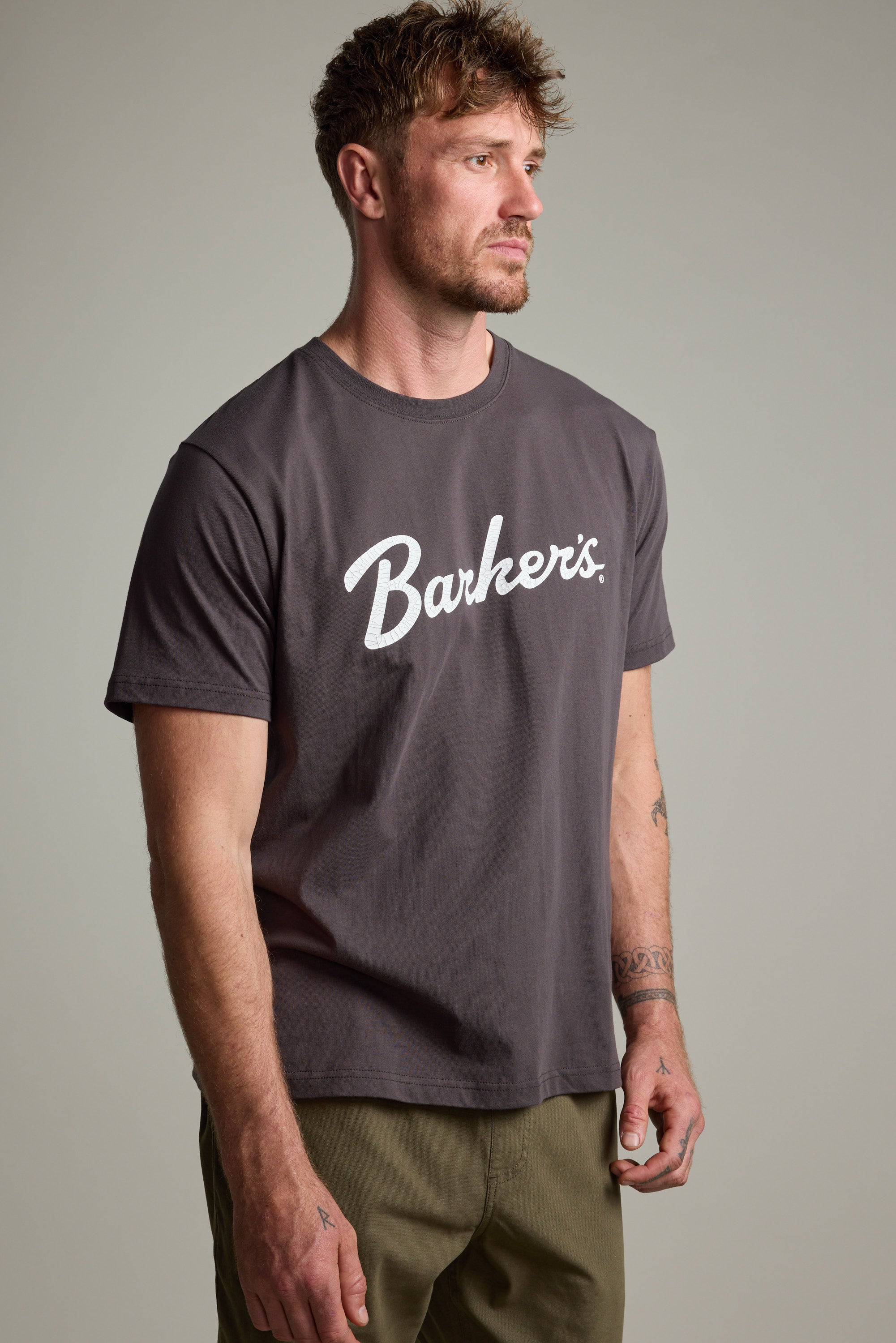 A man with short hair and a beard wears the Barkers Finely Tuned Print Tee, a dark grey organic cotton t-shirt with white script on the chest. He stands facing slightly left, expression neutral, against a plain background.