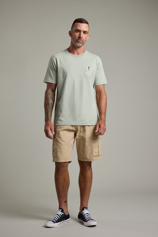 A man with short hair and a trimmed beard faces the camera, wearing a Barkers BMC Sound Sessions Print Tee in light green, beige cargo shorts, and black sneakers. He has tattoos on one arm and stands against a plain gray background with a neutral expression.