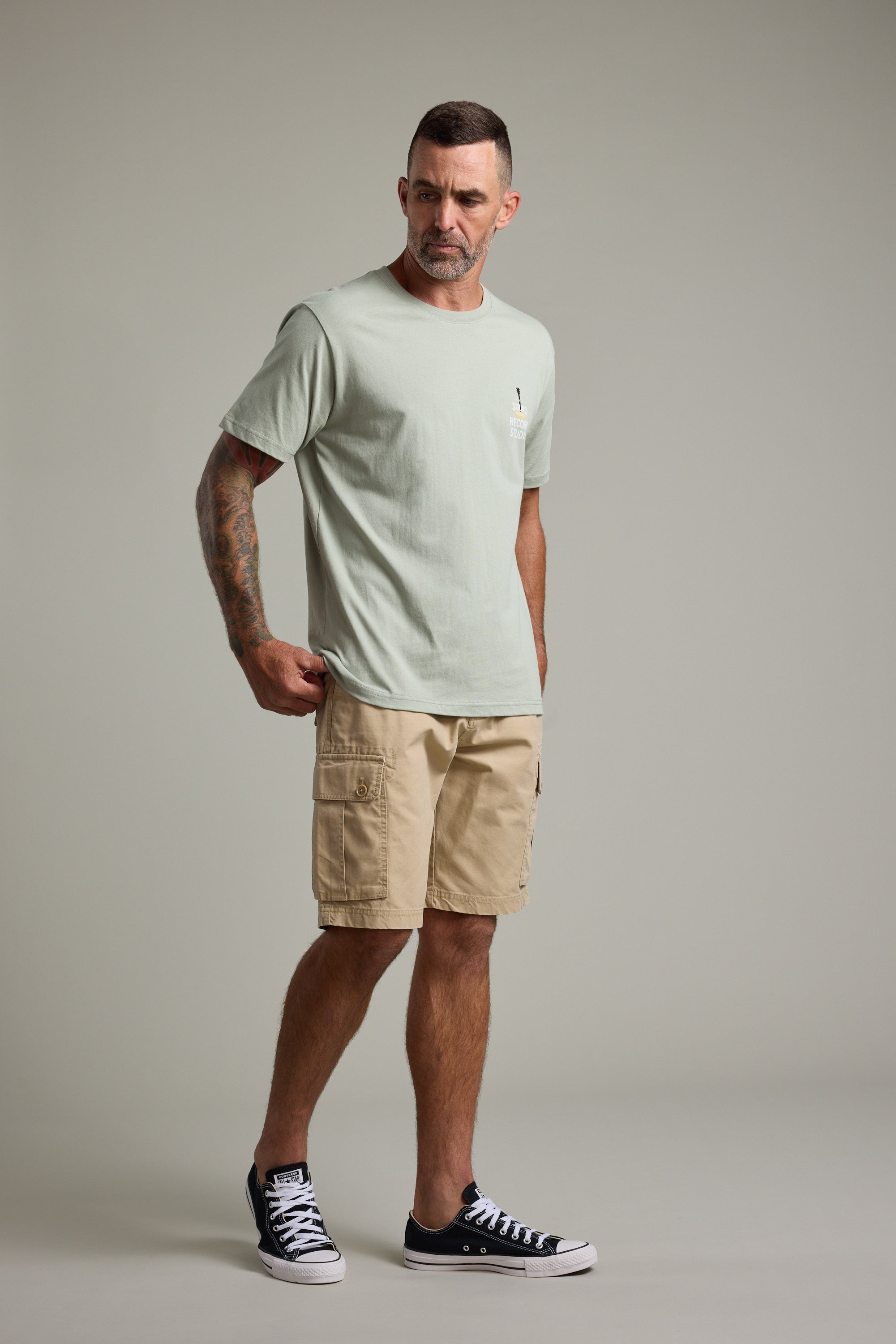 A man with short hair and tattoos wears the Barkers BMC Sound Sessions Print Tee, light green in color, paired with beige cargo shorts and black sneakers. He stands against a gray background, looking left with one hand in his pocket.