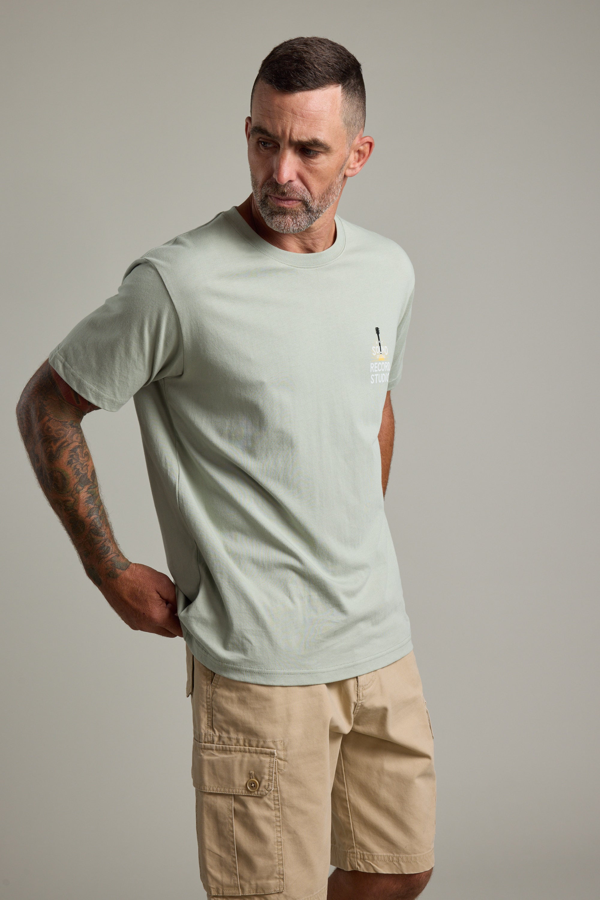 A man with short dark hair and a beard wears a Barkers BMC Sound Sessions Print Tee in light sage green and beige cargo shorts. He stands with hands on hips, looking left against a plain light gray background.