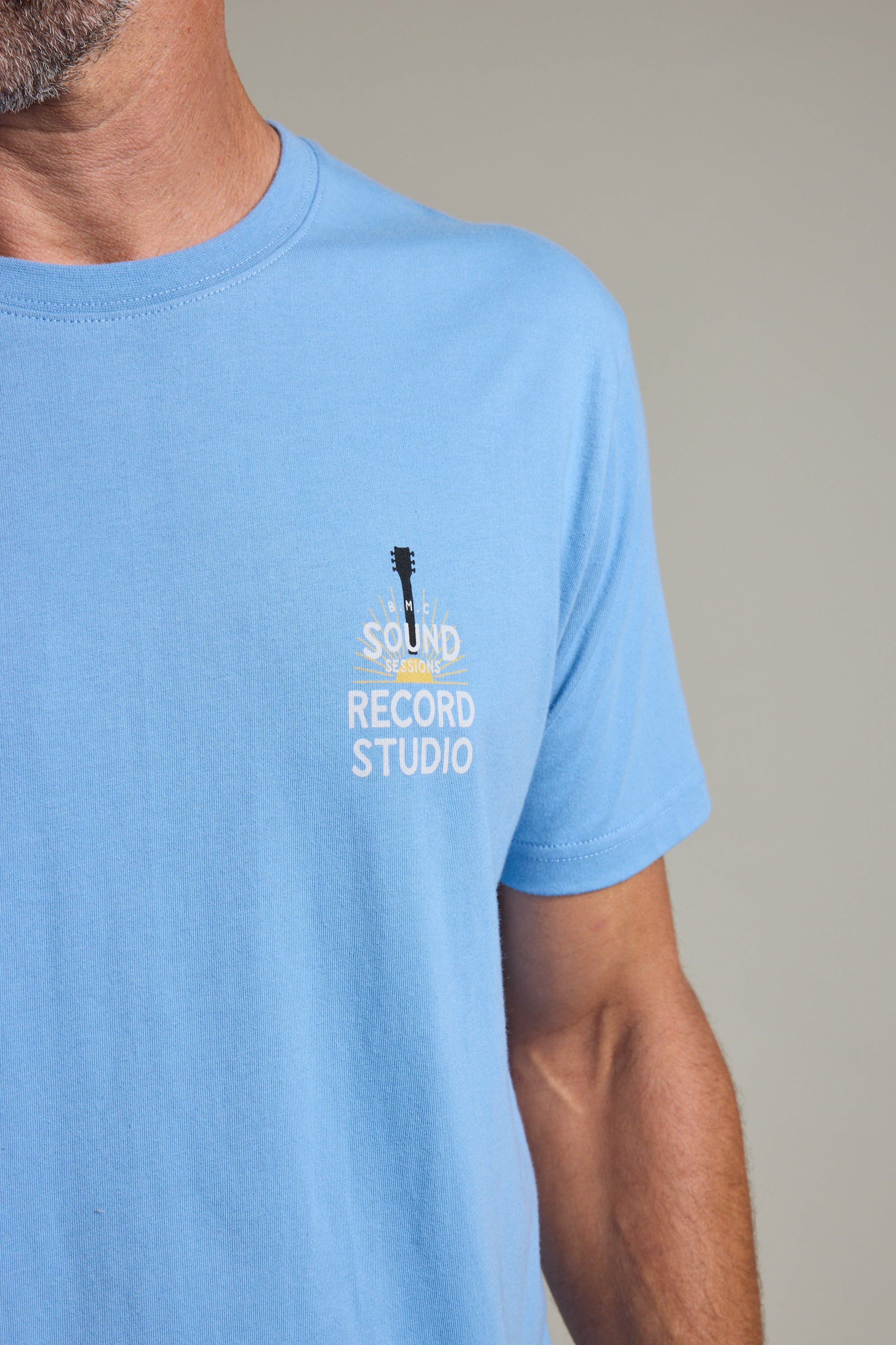 A person wearing a light blue Barkers BMC Sound Sessions Print Tee, featuring a Sound Record Studio guitar logo on the left chest. Only the lower half of the face, neck, and upper torso are visible.