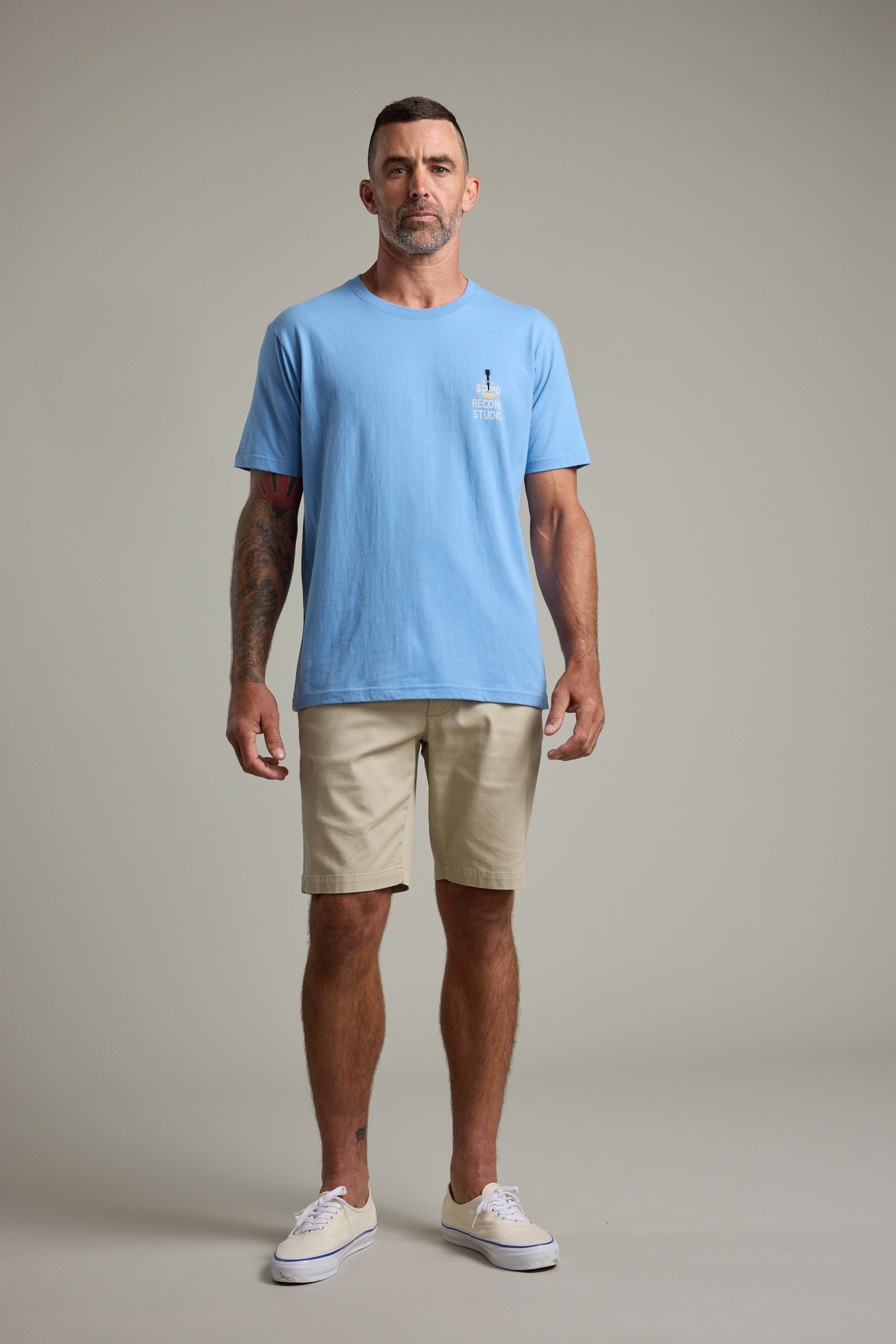 A man with short hair and visible arm tattoos stands facing forward against a plain background, wearing the Barkers BMC Sound Sessions Print Tee in light blue, beige shorts, and white sneakers. He has a neutral expression.