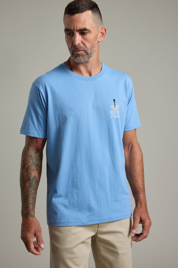 A man with short dark hair and a beard wears the Barkers BMC Sound Sessions Print Tee with beige regular fit pants, standing against a plain gray background. His right arm is tattooed, and he looks slightly down and to his left.