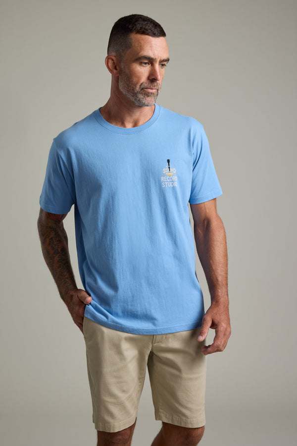 A man with short dark hair and a beard wears a Barkers BMC Sound Sessions Print Tee, light blue in color, and beige shorts, posing with one hand in his pocket against a plain light gray background.