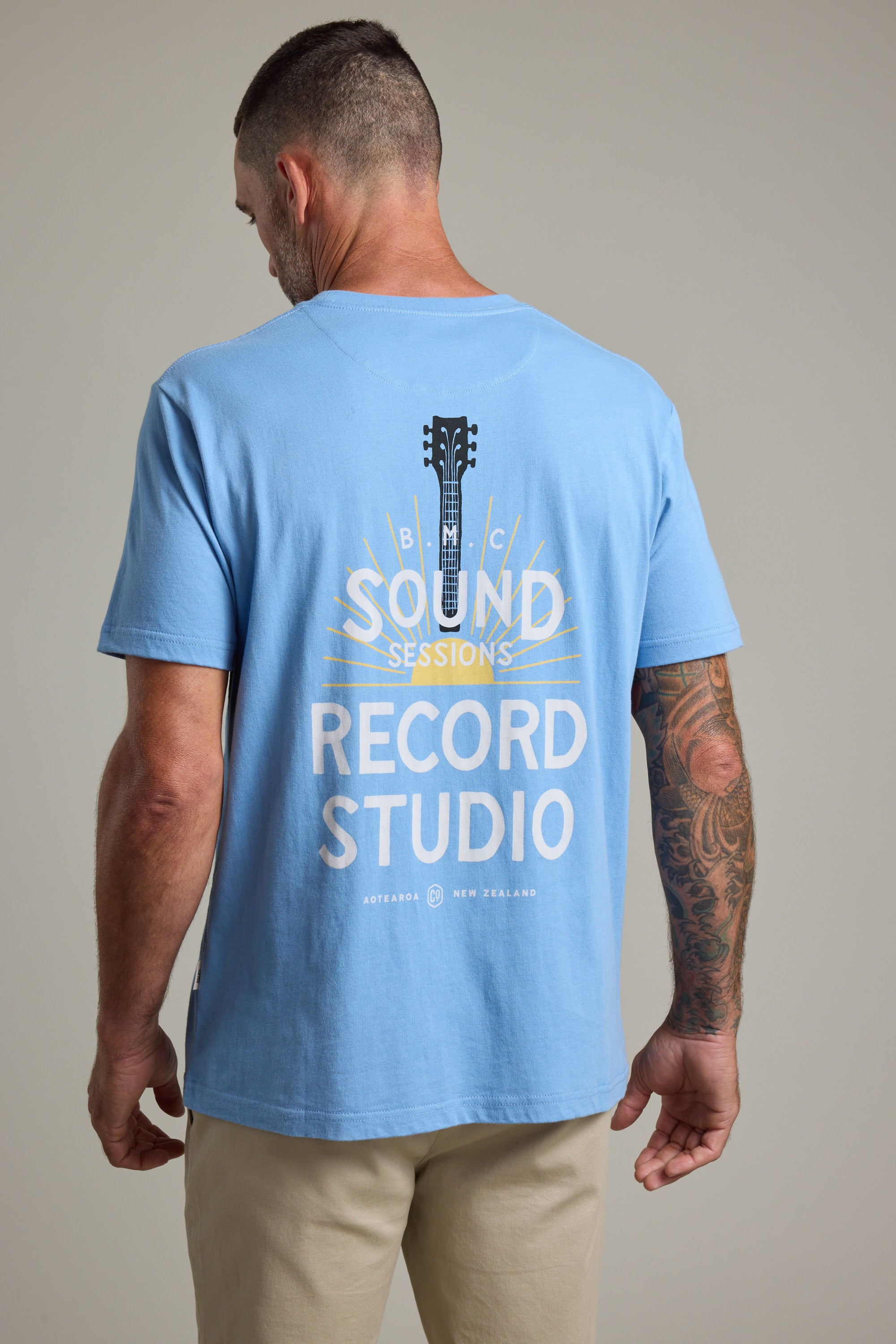 A man with short hair and a tattooed right arm wears the Barkers BMC Sound Sessions Print Tee, a regular fit light blue organic cotton shirt with a guitar graphic and "Sound Sessions Record Studio" on the back, evoking vintage band merch.