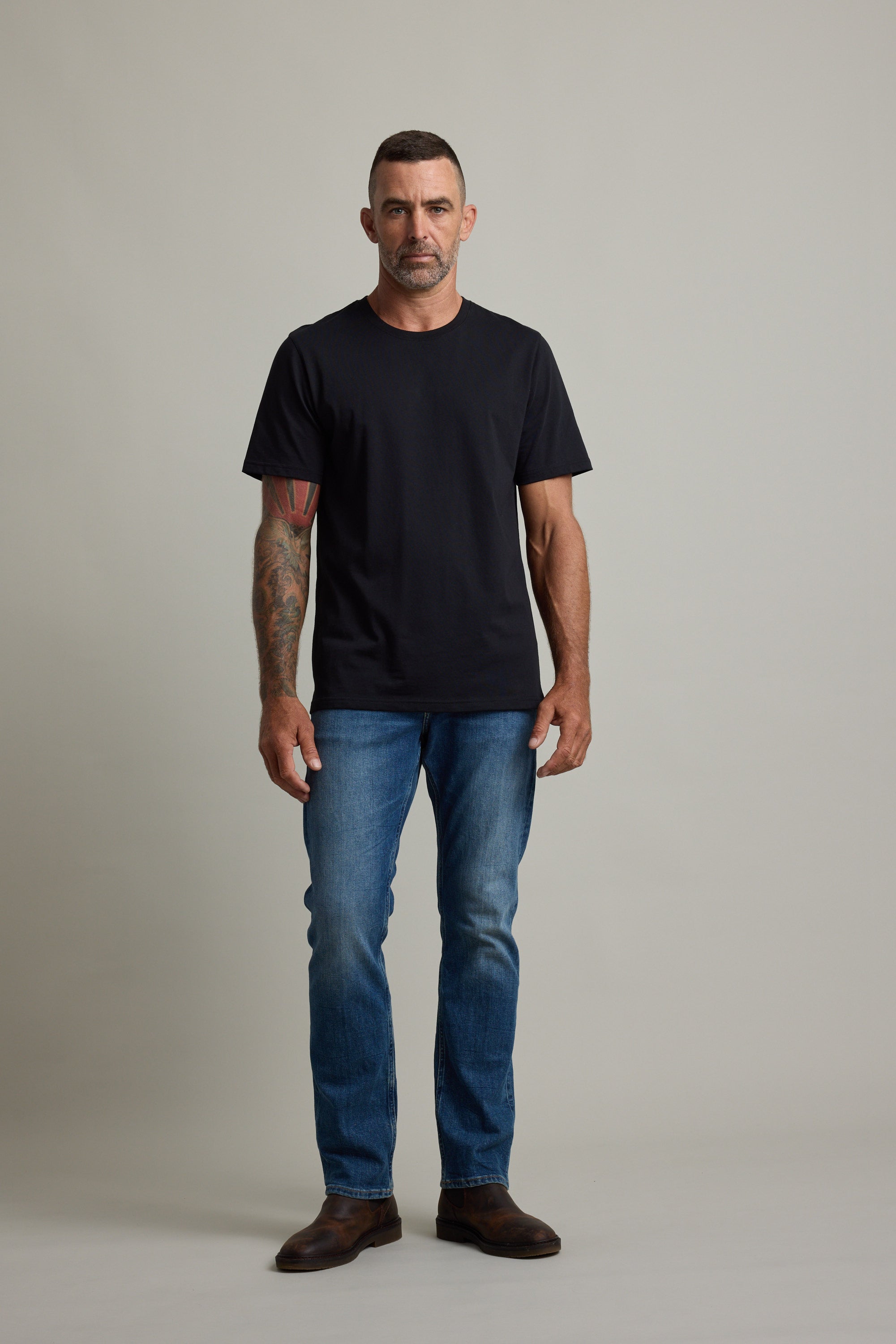 Man in Barkers Good Earth Cotton Essential Tee and blue jeans stands against a plain background.
