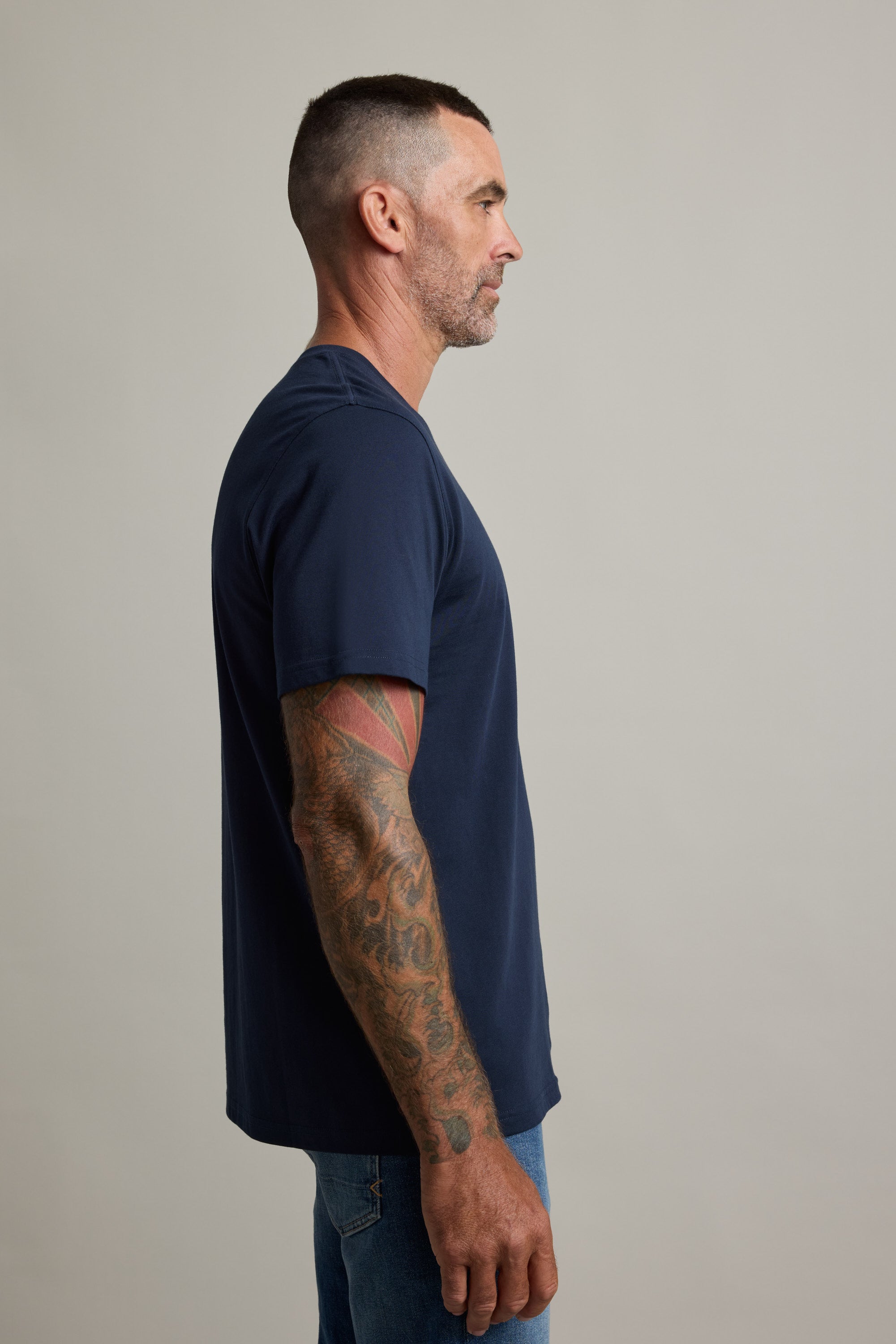 Tattooed man in Barkers Good Earth Cotton Essential Tee, standing in profile against plain background.
