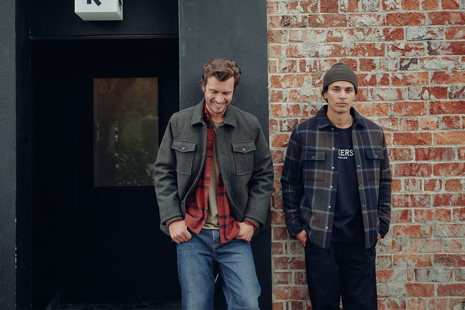 Two men stand side by side against a brick wall. The man on the left is smiling, wearing a green jacket over a red shirt and jeans. The man on the right is wearing a plaid shirt, dark pants, and a beanie, with a neutral expression.