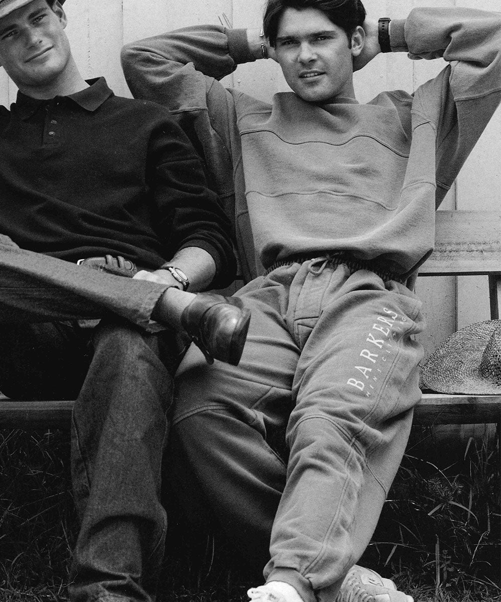 Two young men sit on a bench outdoors. One wears a dark sweater and polo, sitting cross-legged. The other rests his arms behind his head, wearing a light sweatshirt and sweatpants that read BARYLE. A hat lies on the bench beside them.