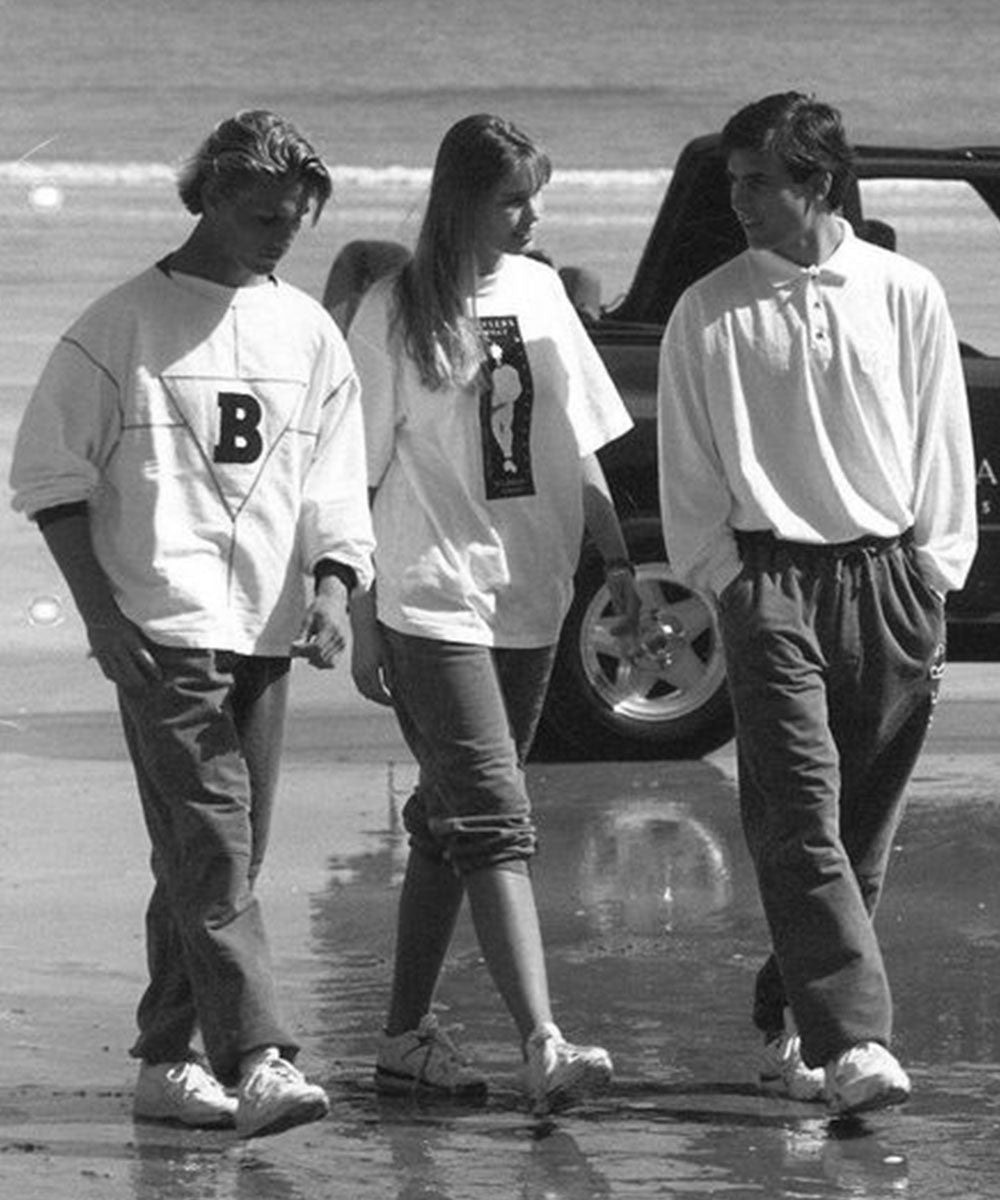 Three young people walk together on a wet beach in casual clothes. Two men wear sweatshirts and rolled-up pants; the woman in the middle wears a T-shirt and shorts. A car and the ocean are seen in the background.