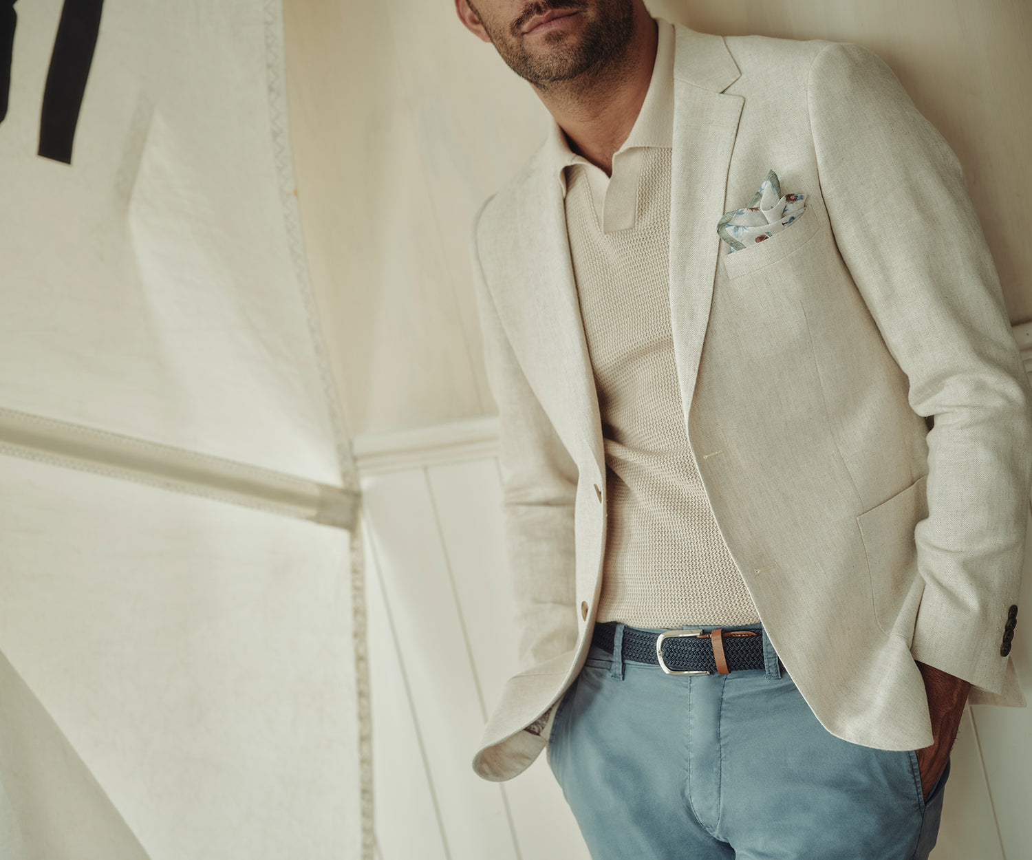 A man dressed in a light beige blazer over a matching knit polo shirt, blue trousers, and a navy belt stands against a cream-colored wall. A printed pocket square is visible in his blazer pocket. His face is partially out of frame.