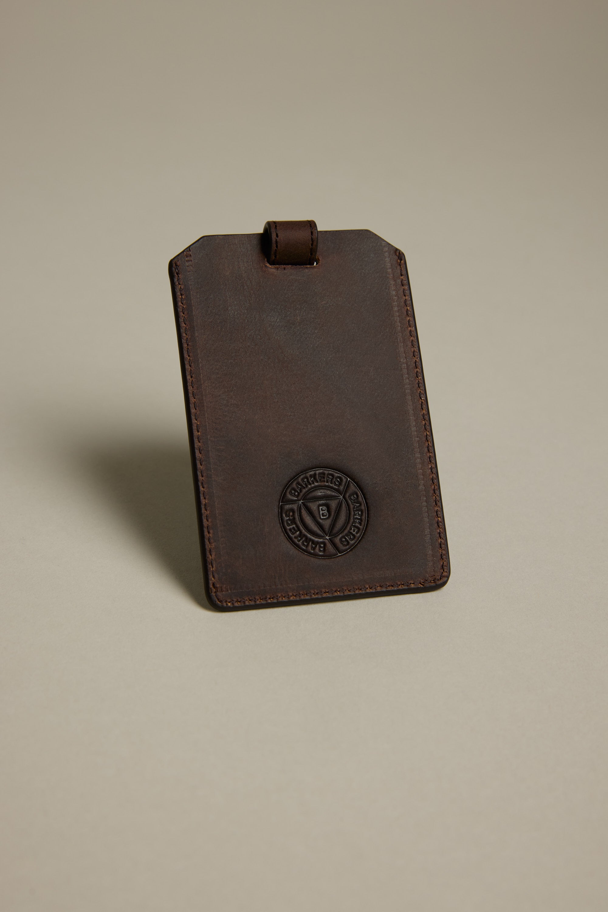 The Barkers Voyage Leather Luggage Tag stands upright against a plain beige background—crafted in brown leather with stitched edges and an embossed circular logo, it’s the perfect corporate gift or stylish travel accessory.