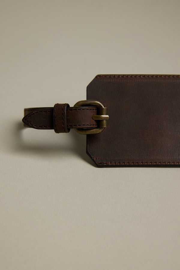 The Barkers Voyage Leather Luggage Tag, shown in brown with a brass buckle and stitched edges, lies flat on a beige surface. Its adjustable strap makes this rectangular travel accessory perfect as a corporate gift.