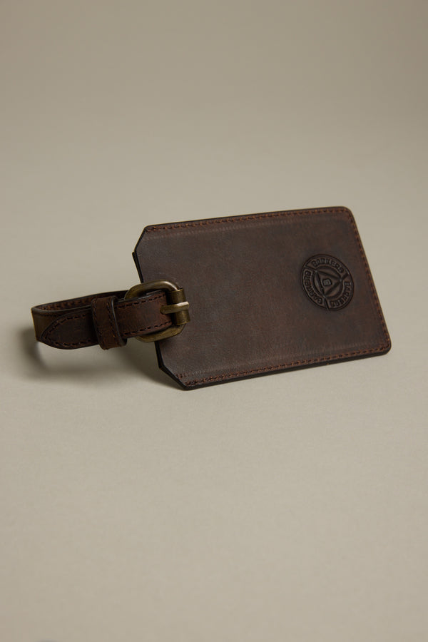 The Barkers Voyage Leather Luggage Tag, featuring a brass buckle and circular embossed logo, rests on a plain beige surface—ideal as a travel accessory or corporate gift.