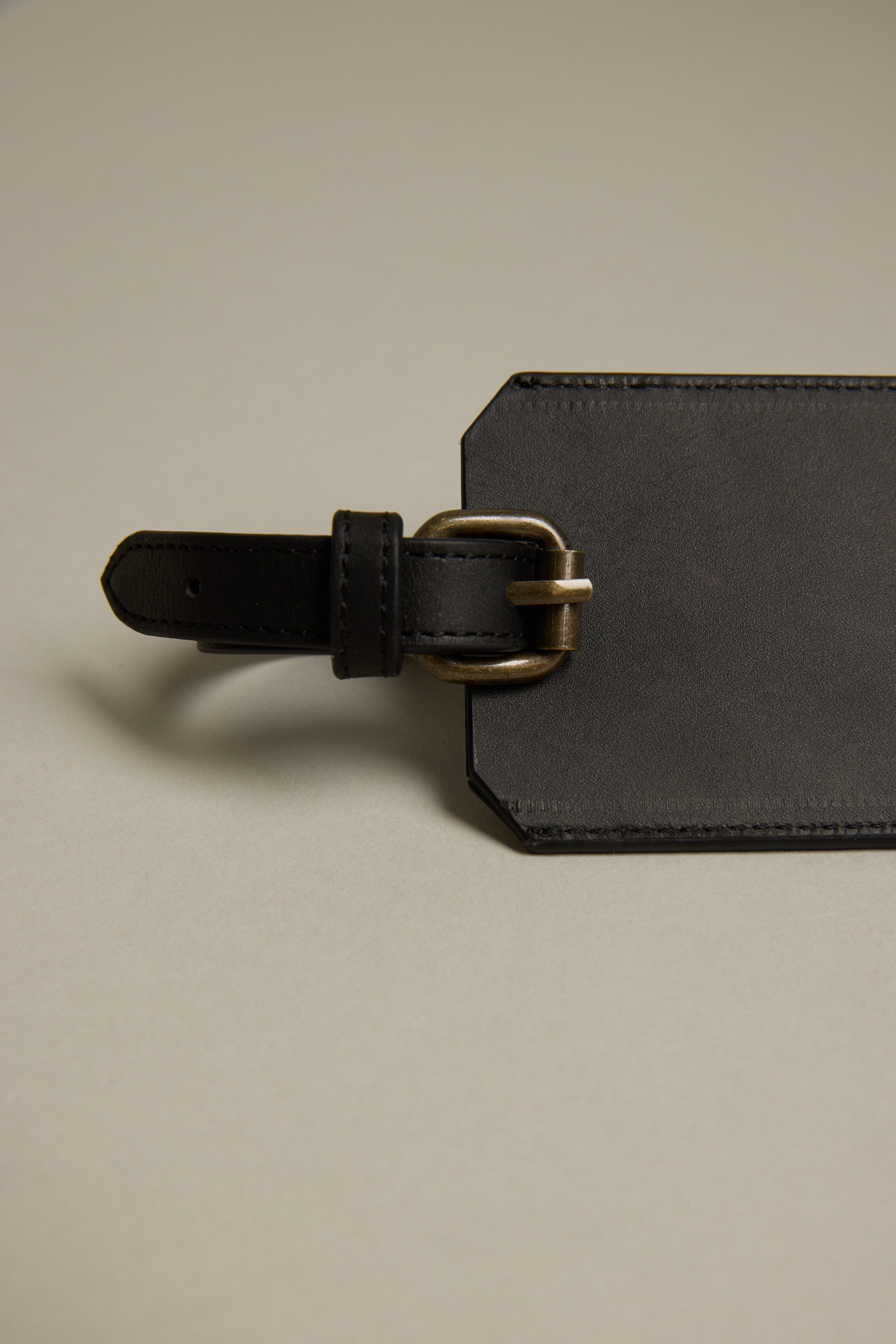 A close-up of the Barkers Voyage Leather Luggage Tag in black, featuring a brass buckle and displayed on a beige surface—a stylish essential for frequent flyers.