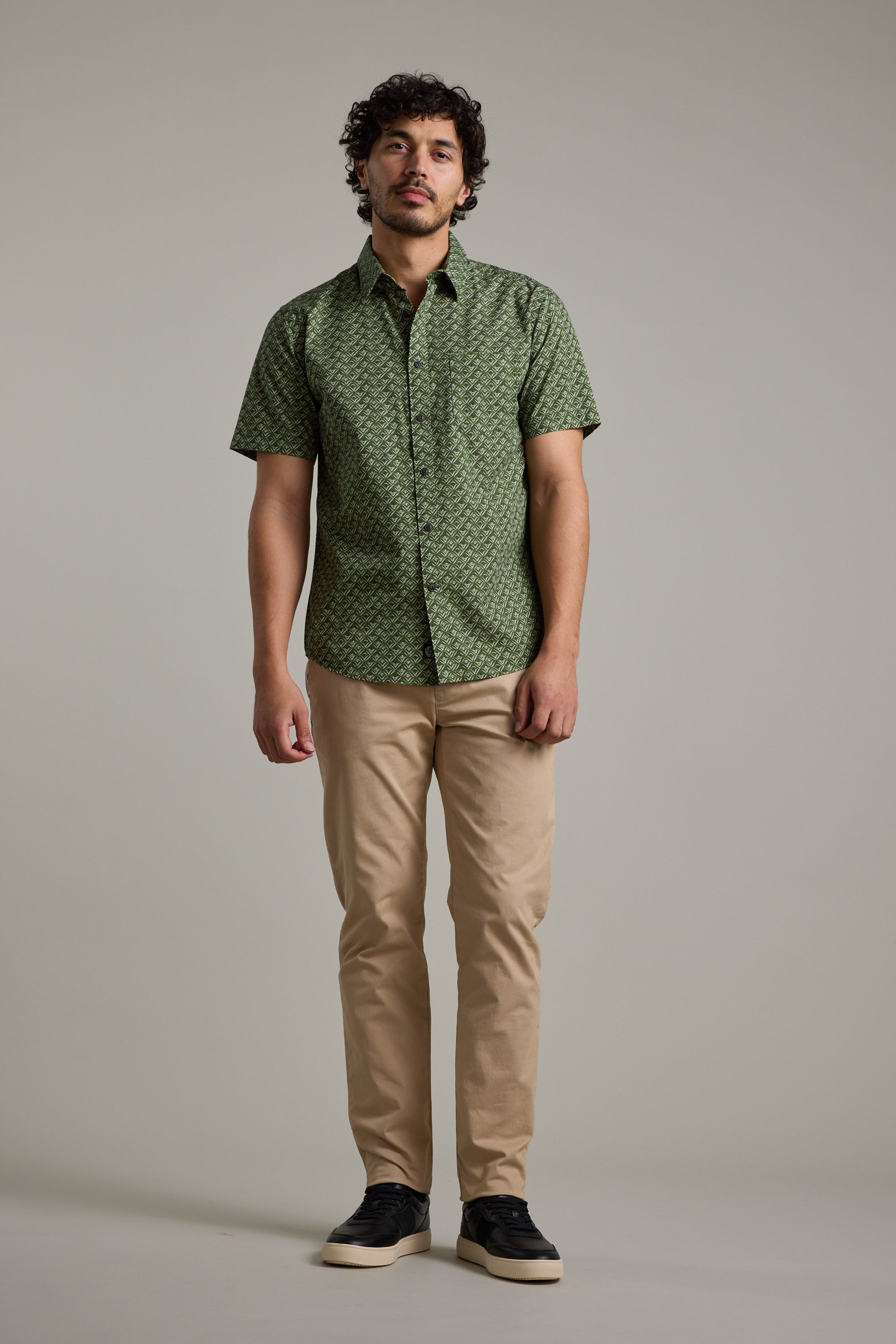 A man stands against a plain background wearing the Barkers Oxnard Print Shirt in green organic cotton, paired with beige pants and black sneakers. He looks forward with a neutral expression and relaxed posture.