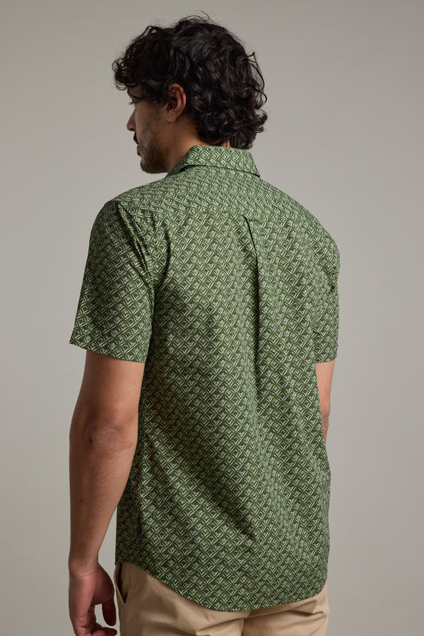 A man with curly dark hair, seen from behind, wears the Barkers Oxnard Print Shirt in green organic cotton and beige pants, standing against a plain gray background.