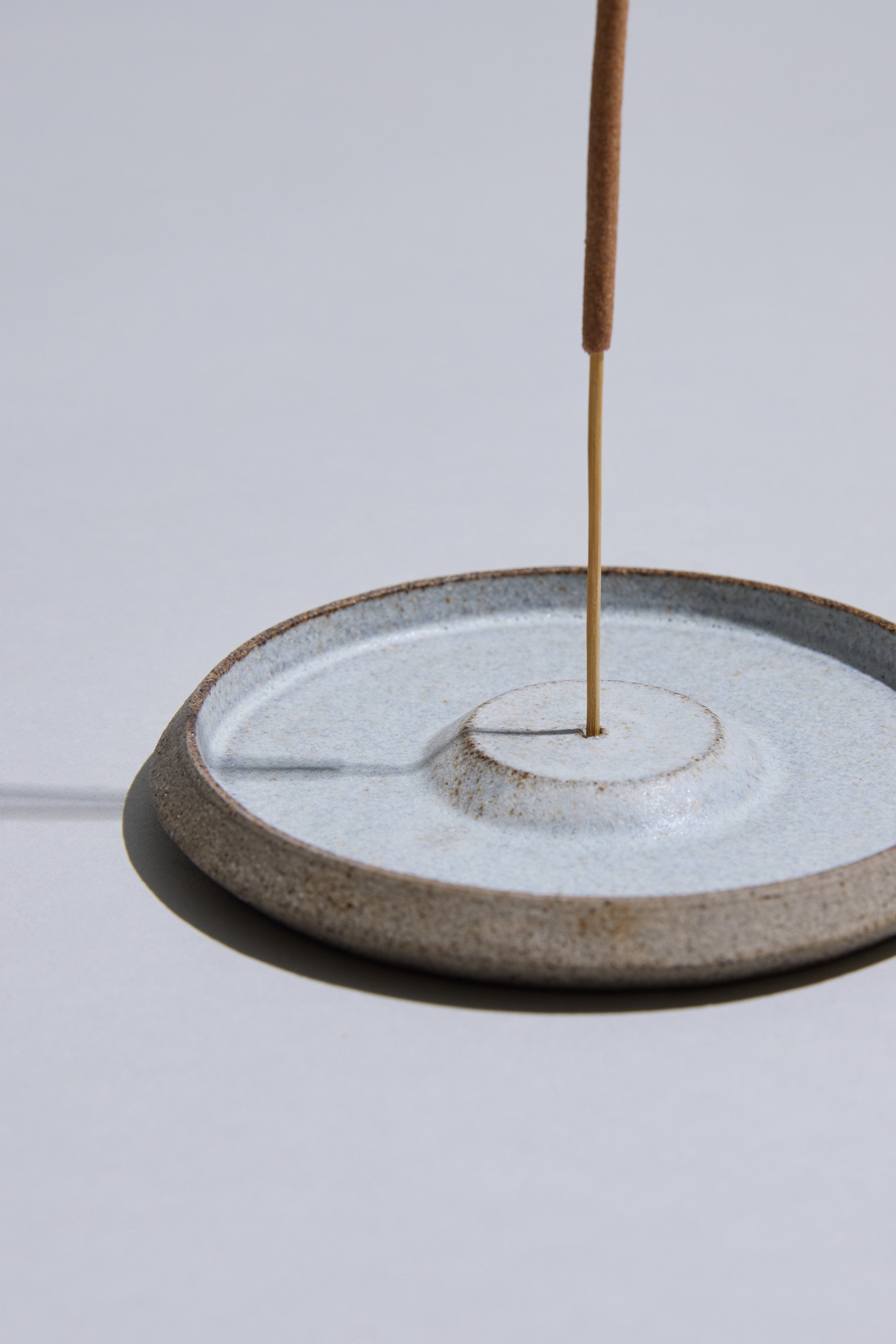 A brown incense stick stands upright in a round, light blue Studio Foon Incense Dish by Other, casting a shadow on a smooth, light-gray surface.