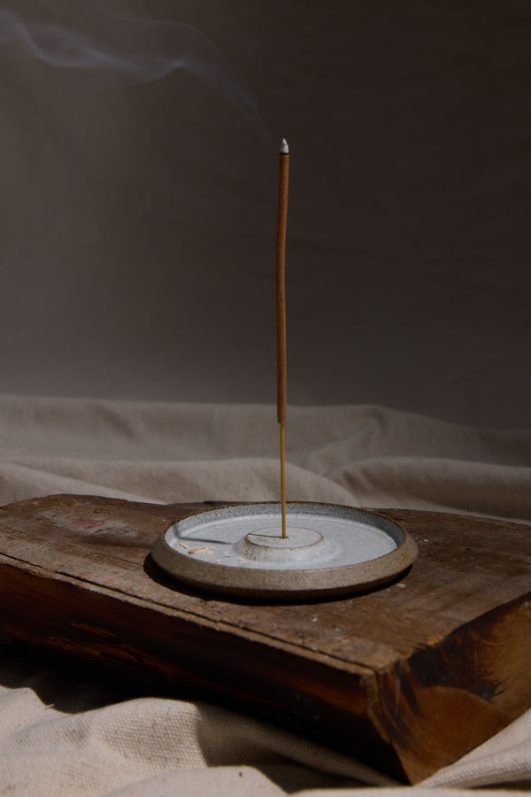 A slender incense stick burns upright in the Studio Foon Incense Dish by Other, crafted from natural New Zealand clay. Wisps of smoke drift upward as the holder sits on a rustic wooden slab against a soft, neutral fabric background.