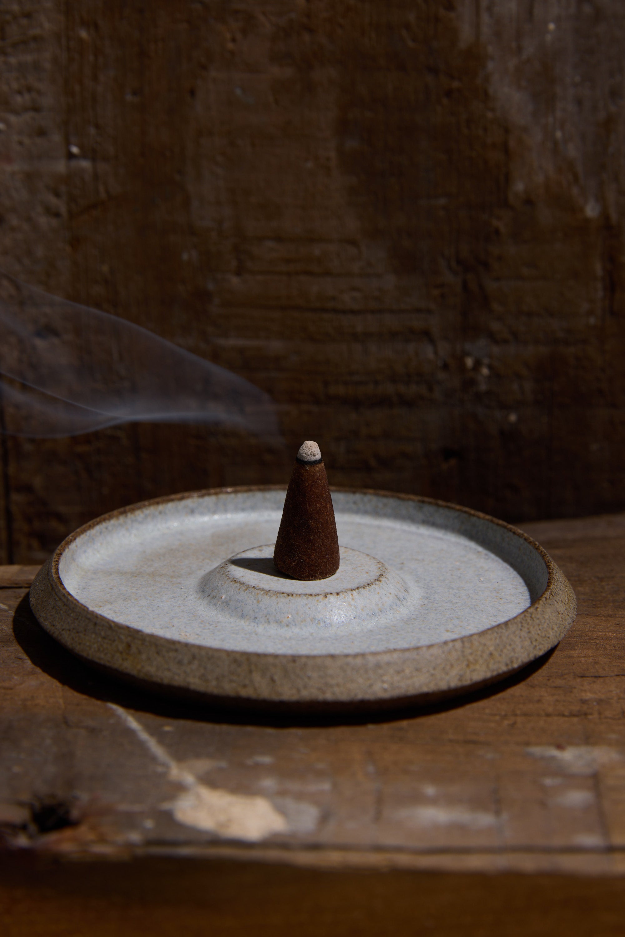 A single incense cone burns on the handcrafted Studio Foon Incense Dish by Other, releasing a delicate wisp of smoke against a rustic wooden background.
