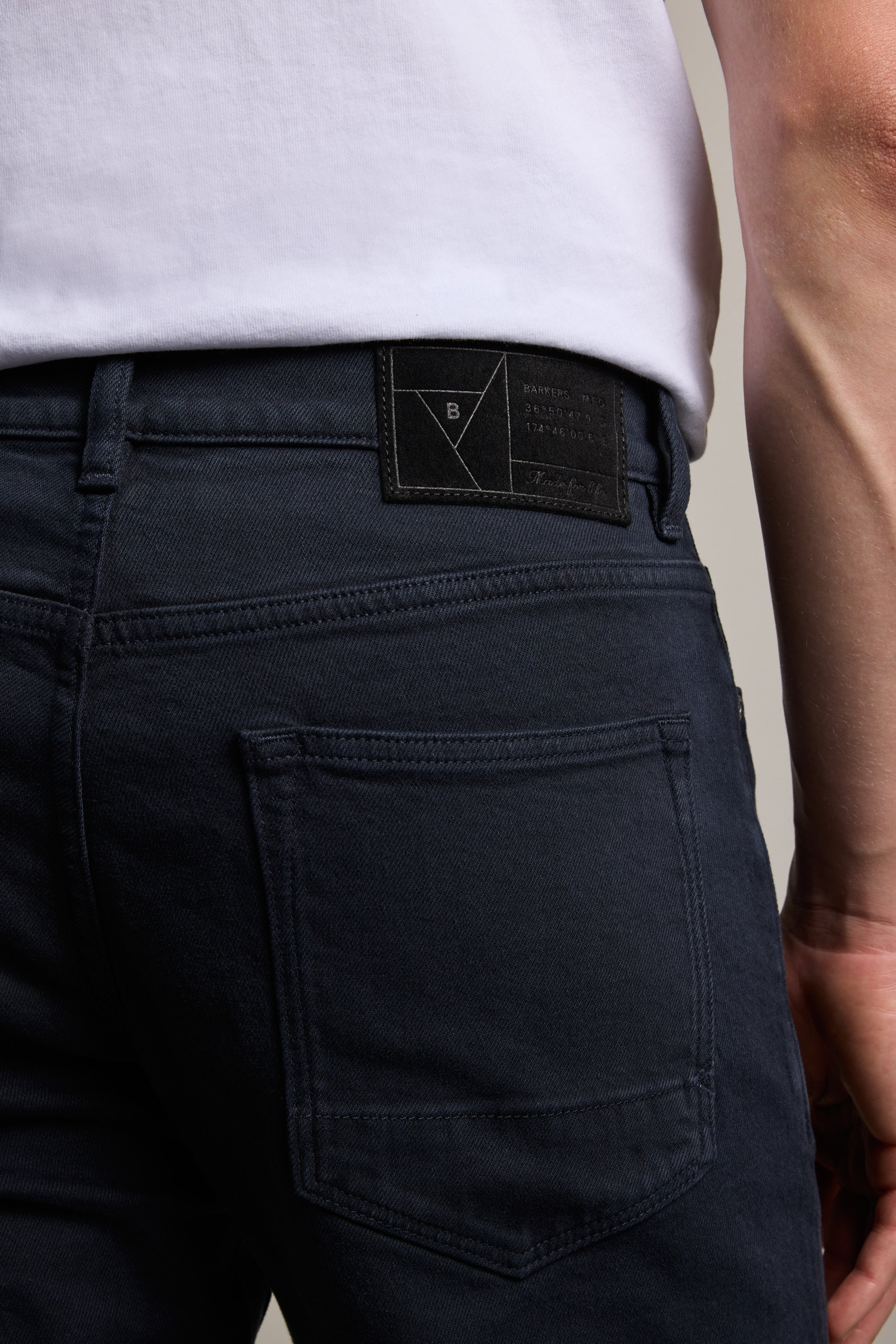 A close-up of someone wearing Barkers Rambler Slim Coloured Jeans in dark blue with a white shirt, highlighting the back pocket, waistband, and the black label on these slim jeans.