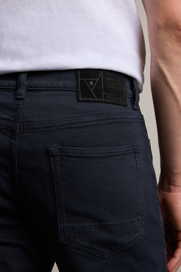 A close-up of someone wearing Barkers Rambler Slim Coloured Jeans in dark blue with a white shirt, highlighting the back pocket, waistband, and the black label on these slim jeans.