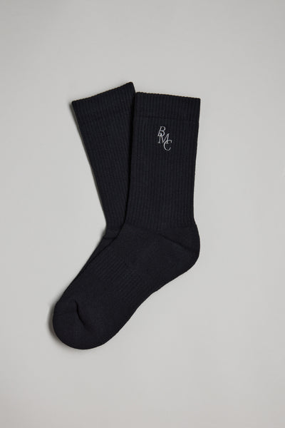 A pair of black Barkers BMC Ribbed Socks is laid flat on a light gray surface, featuring a small white RMC logo embroidered near the top of one sock.