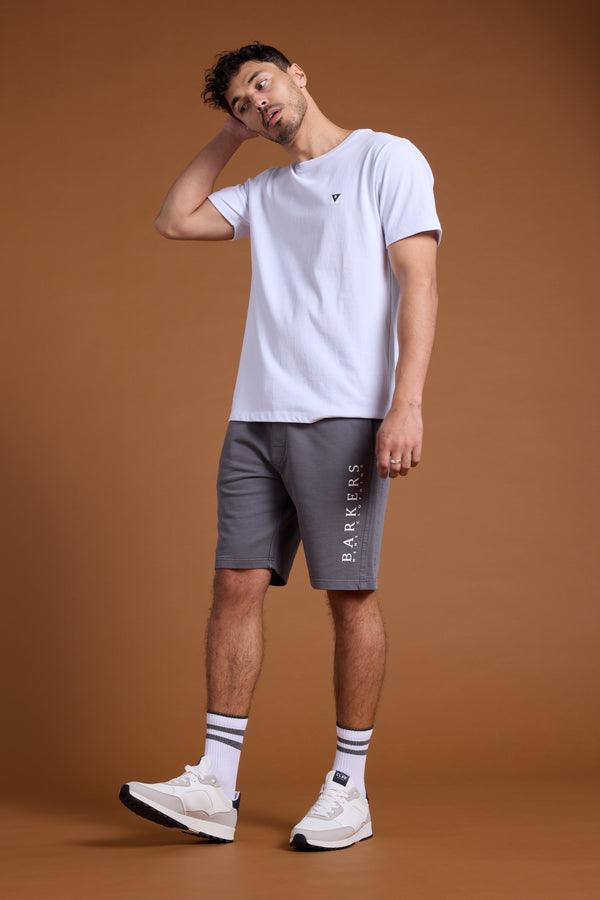 A man stands against a brown background wearing a white tee, Barkers Organic Classic Print Track Short with “BARRIERS” on the side, striped white socks, and white sneakers. He tilts his head and has one hand behind his neck.