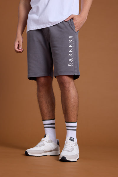 A person stands against a plain brown background wearing a white T-shirt, Barkers Organic Classic Print Track Short in grey with the heritage logo, white socks with black stripes, and white sneakers. The upper face and head are not visible.