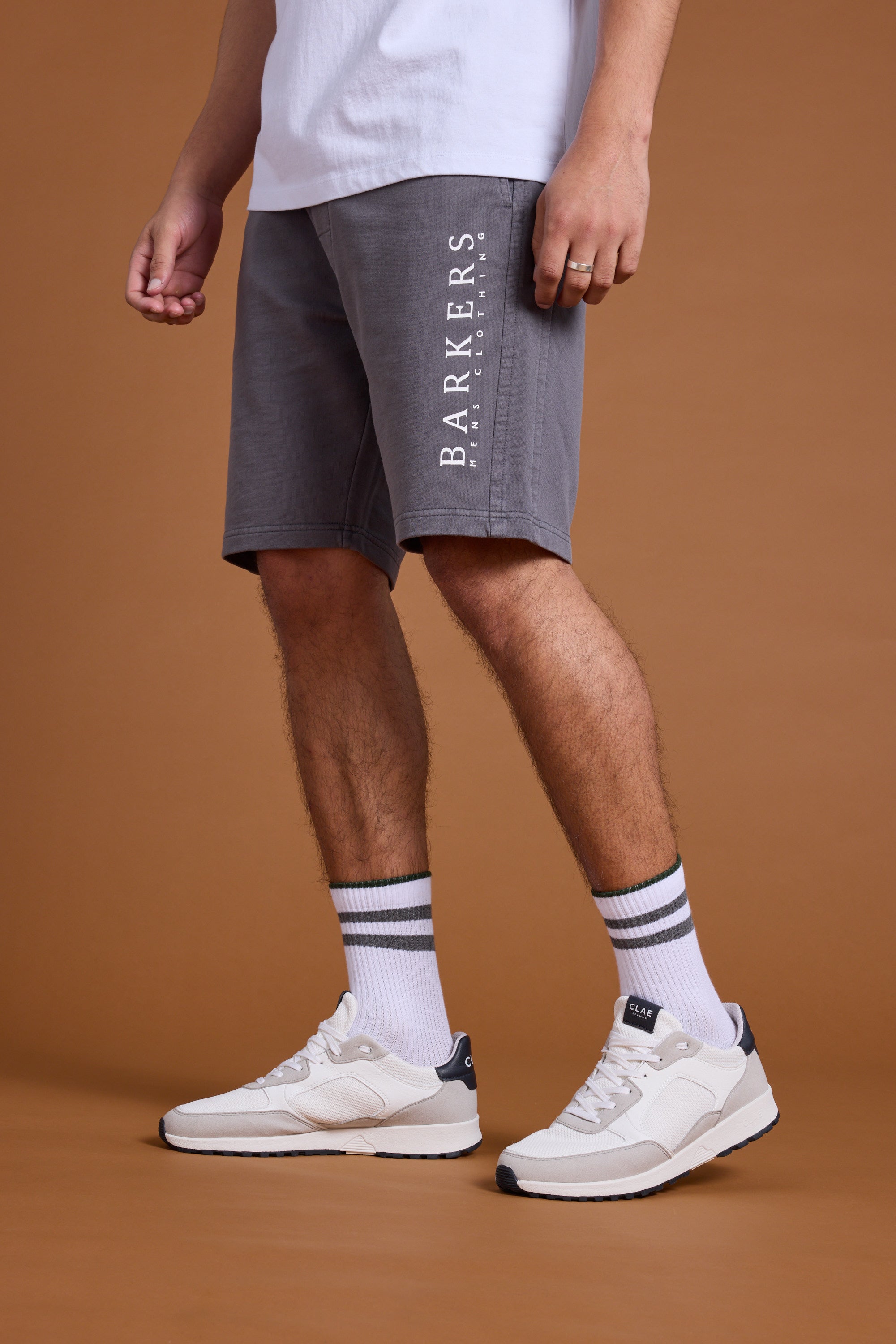 A person wears Barkers Organic Classic Print Track Shorts in grey with the heritage logo, a white T-shirt, white socks with navy stripes, and white sneakers, standing against a plain brown background. Only the lower body is visible.
