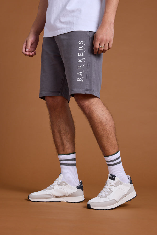 A person wears Barkers Organic Classic Print Track Shorts in grey with the heritage logo, a white T-shirt, white socks with navy stripes, and white sneakers, standing against a plain brown background. Only the lower body is visible.